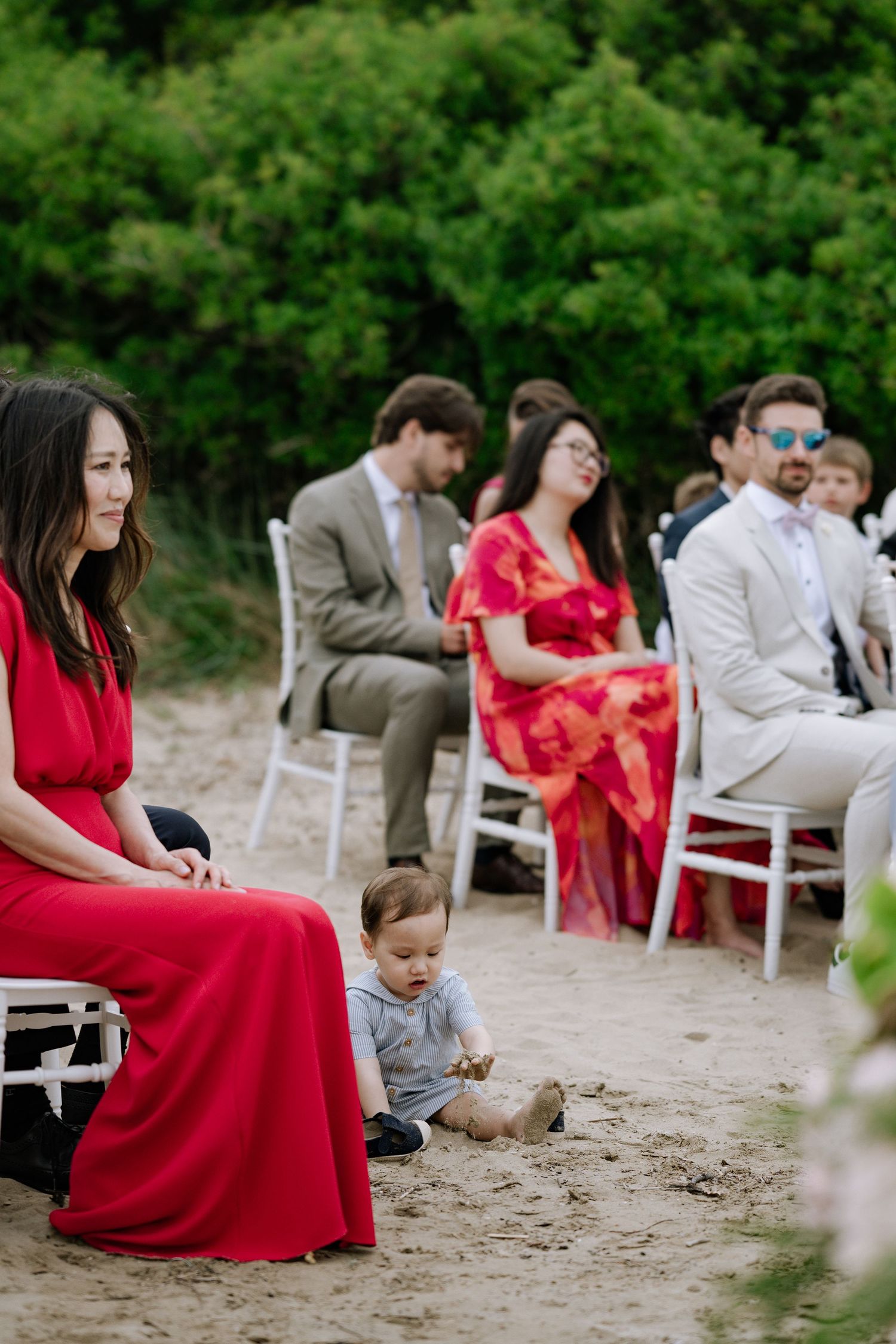 Wedding guests seated in white chairs on sandy beach ceremony with baby playing in sand foreground.
