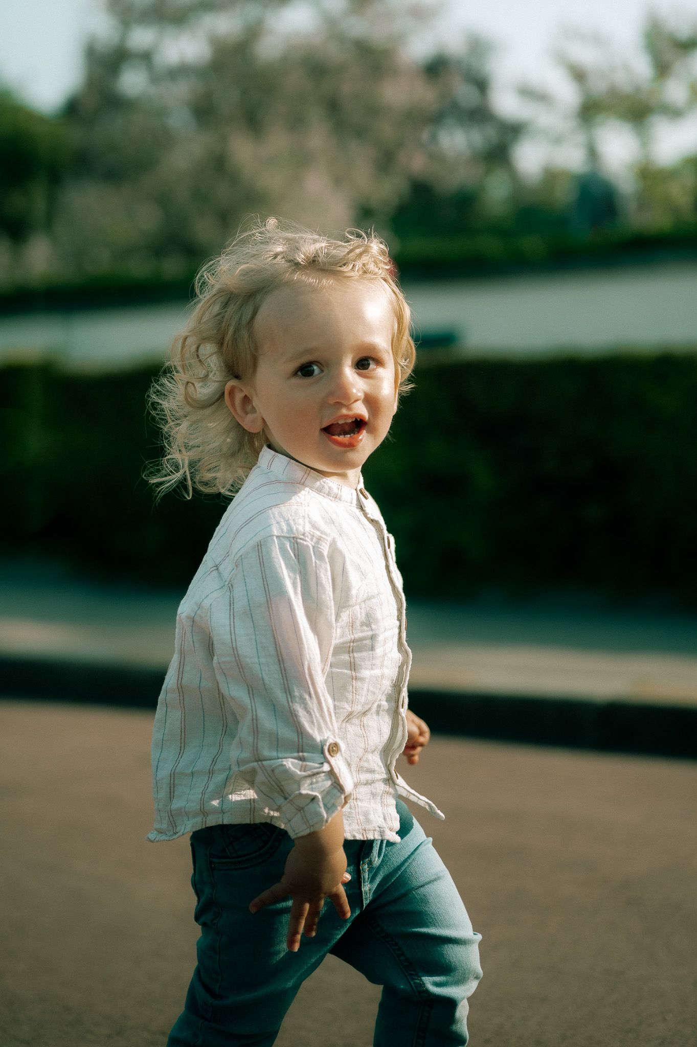 Adorable curly-haired toddler in a striped white shirt and jeans walking outdoors with a joyful expression.