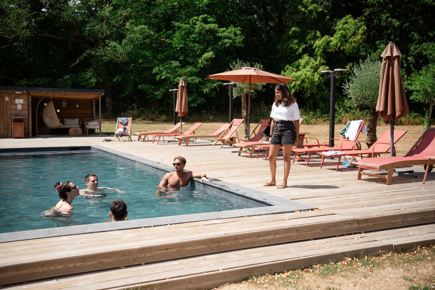 Guests swimming and relaxing by an outdoor pool surrounded by lounge chairs and umbrellas in a lush green setting.