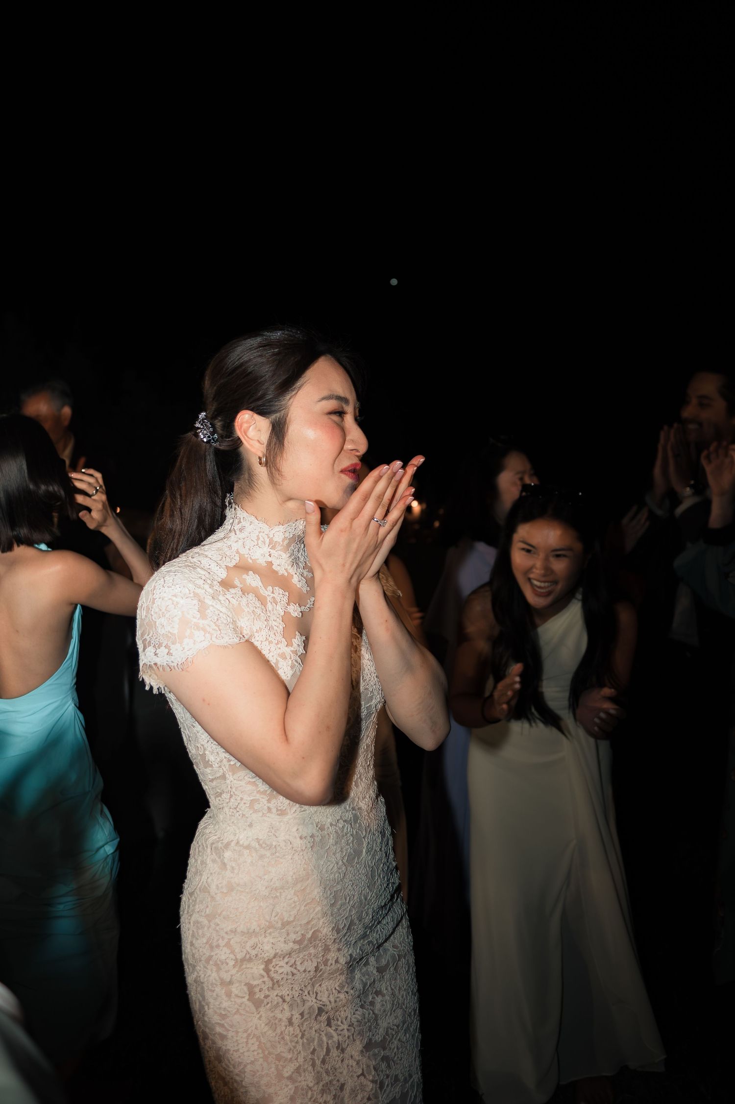 Bride in lace dress claps with joy while dancing with guests at her nighttime wedding celebration.