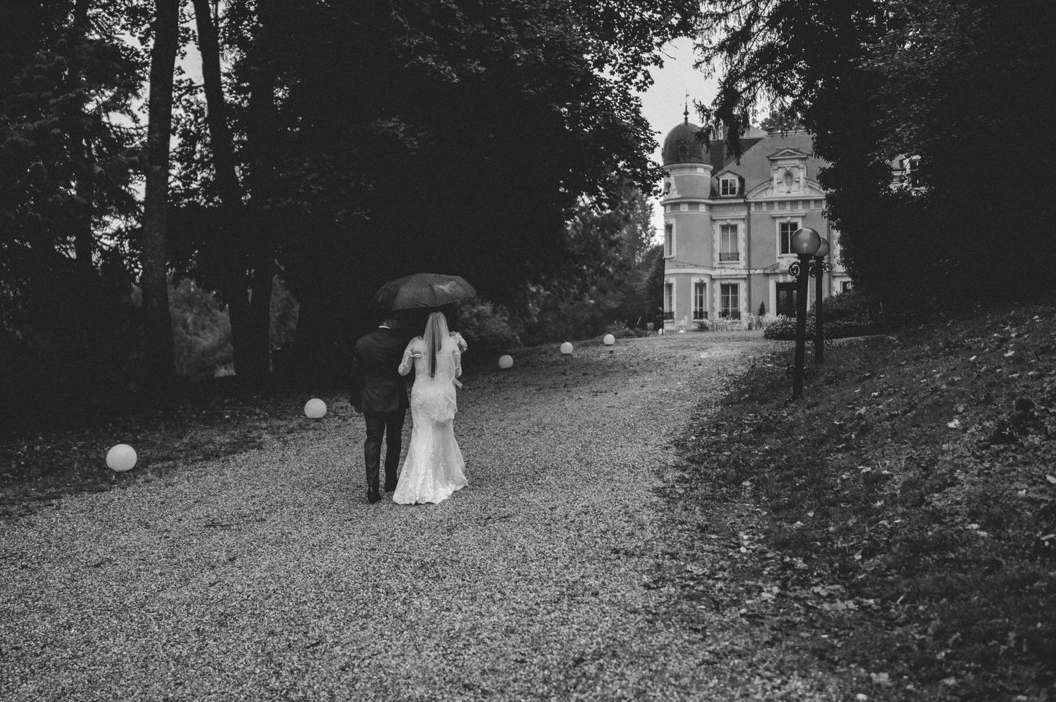 Black and white photo of bride and groom walking under umbrella on rainy day toward chateau.