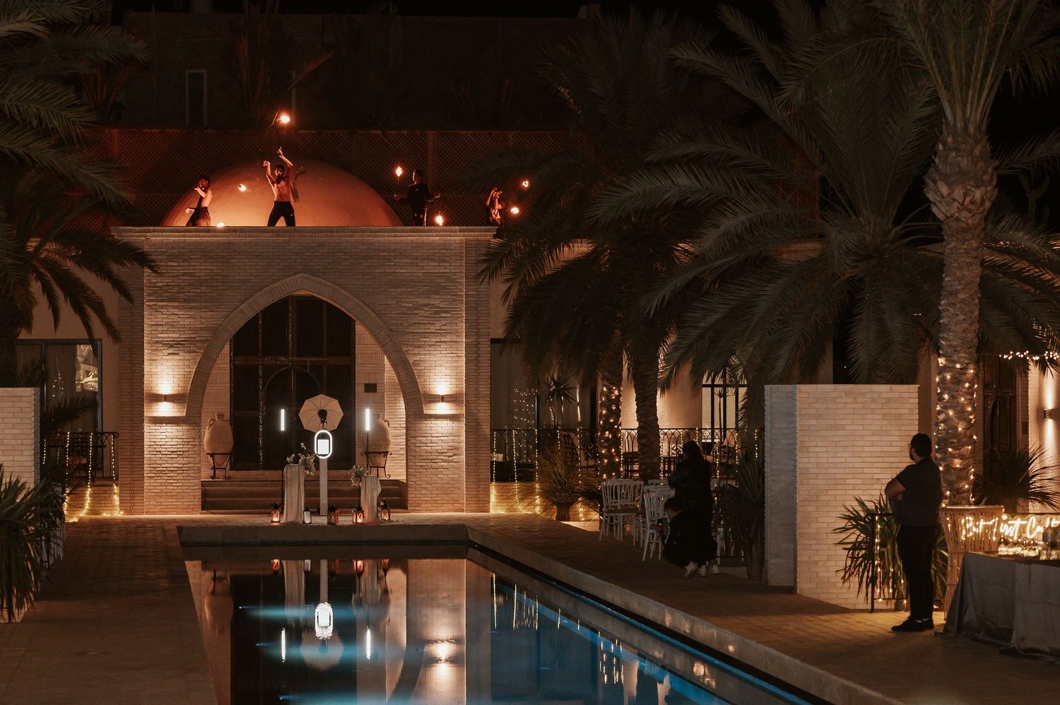 Elegant outdoor wedding venue with pool, palm trees, and warm architectural lighting at night.