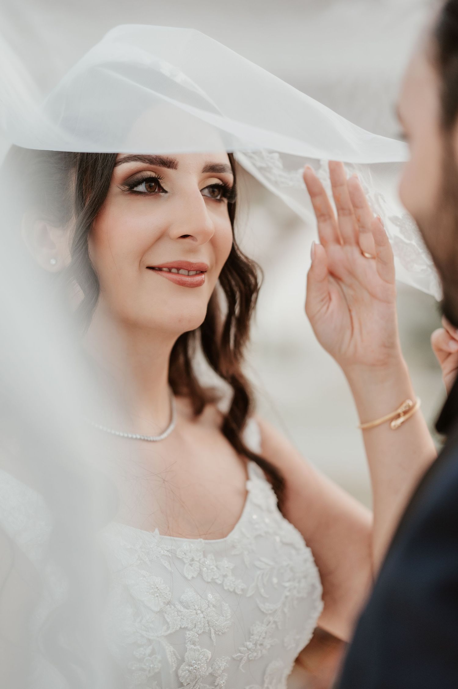 Groom lifts bride's veil to reveal her elegant bridal makeup and pearl necklace on wedding day.