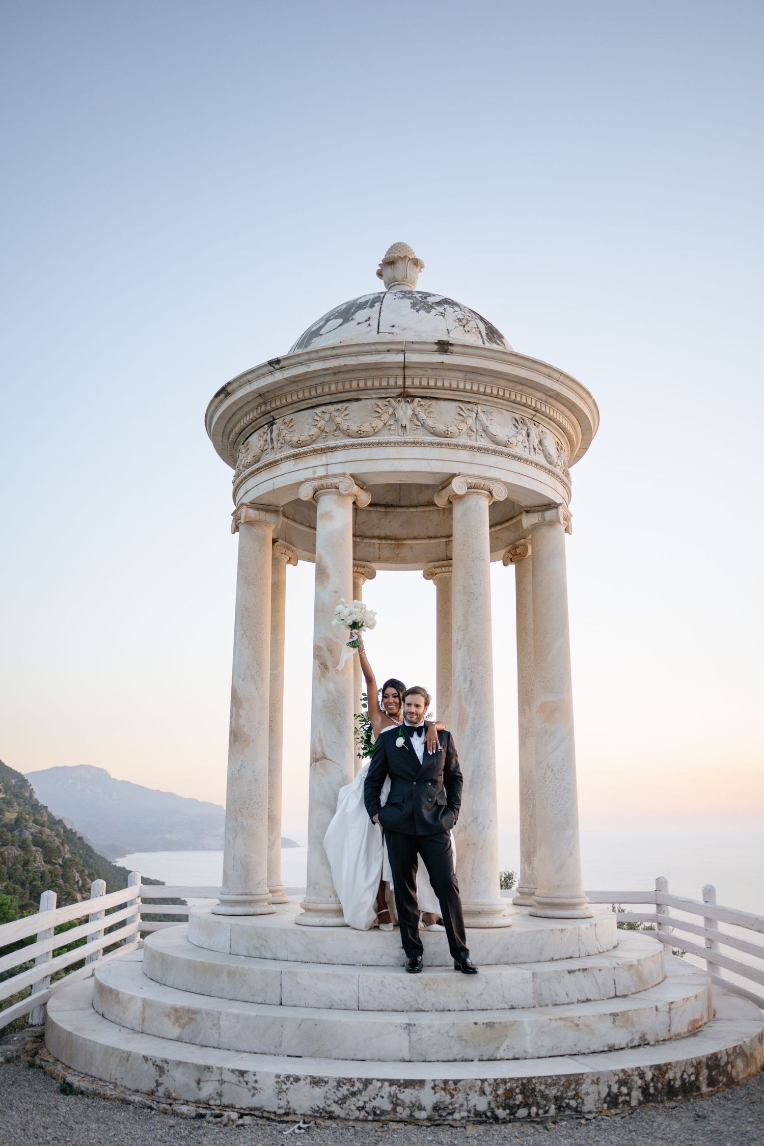 Joyful bride and groom celebrate atop marble rotunda steps with bouquet raised against clear sky.