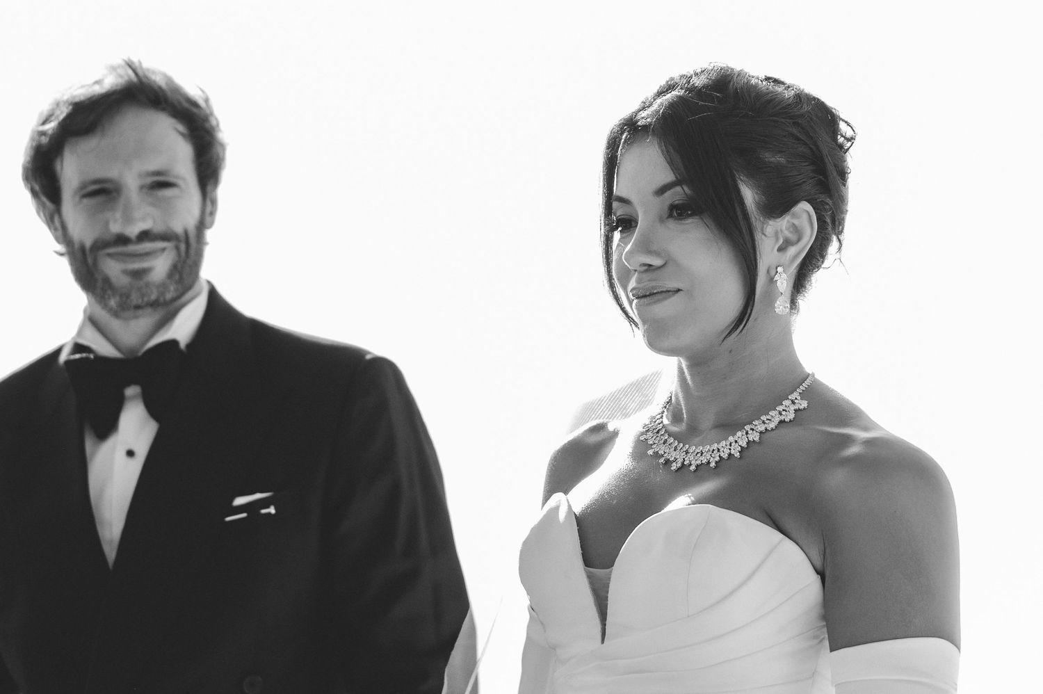 Black and white close-up of a smiling groom and bride wearing a diamond necklace at the altar.