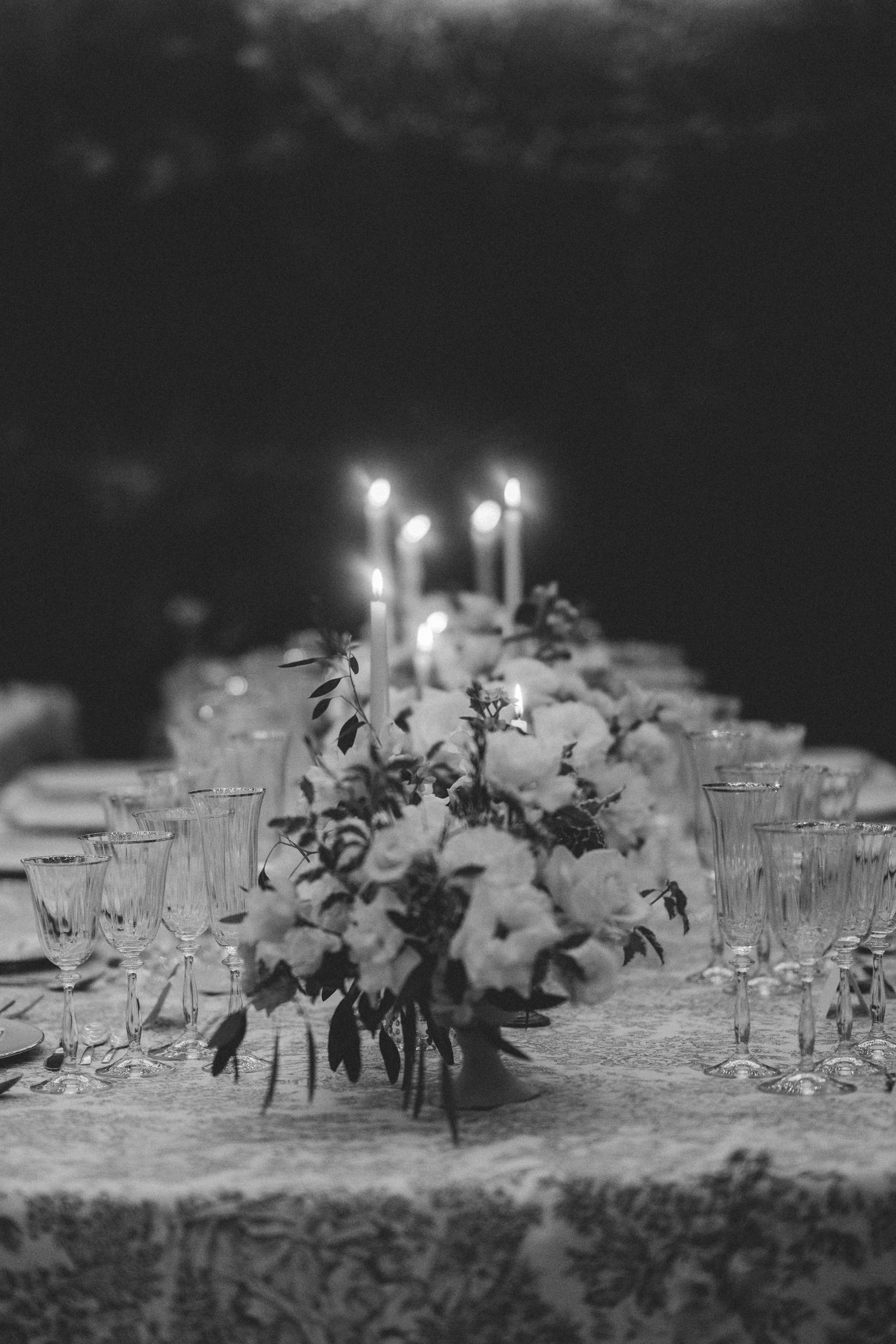 Black and white photo of romantic wedding table with lit taper candles and floral centerpiece on patterned linen.