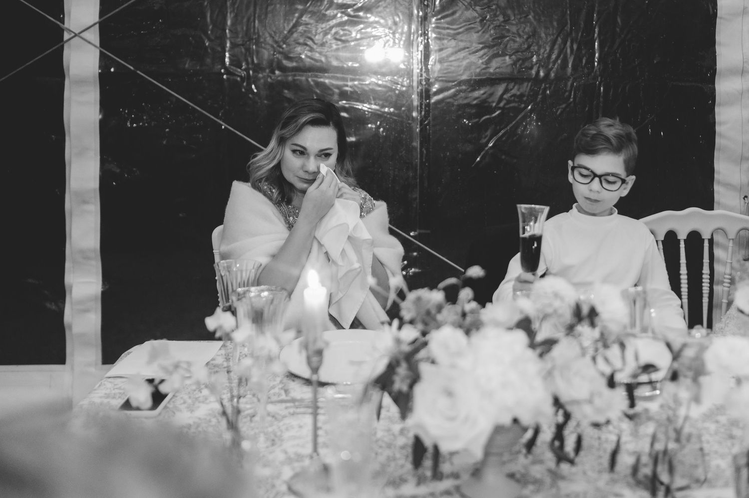 Black and white photo of an emotional wedding guest wiping tears at a candlelit reception table.
