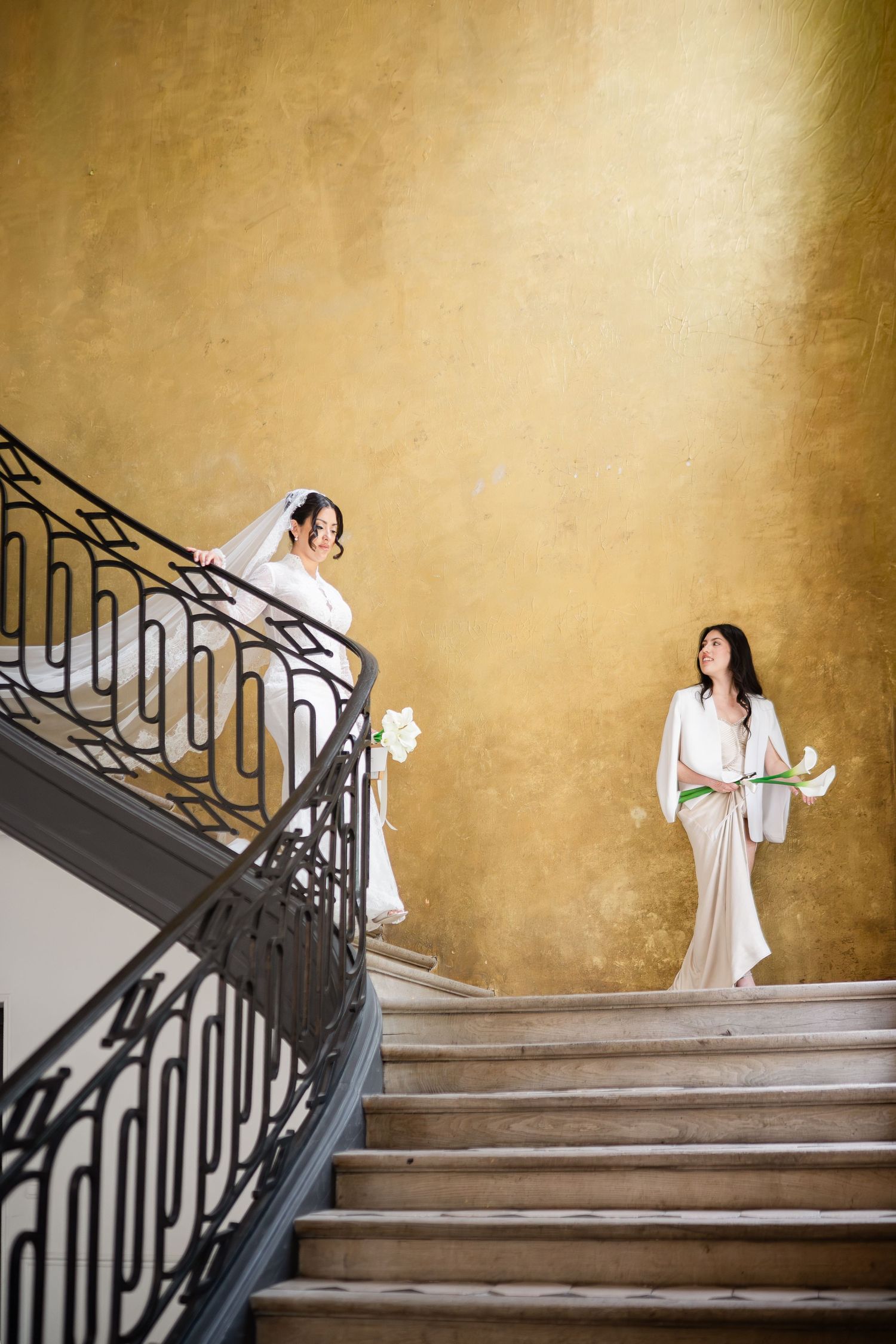 Two brides in white gowns holding bouquets on an elegant staircase with golden walls.