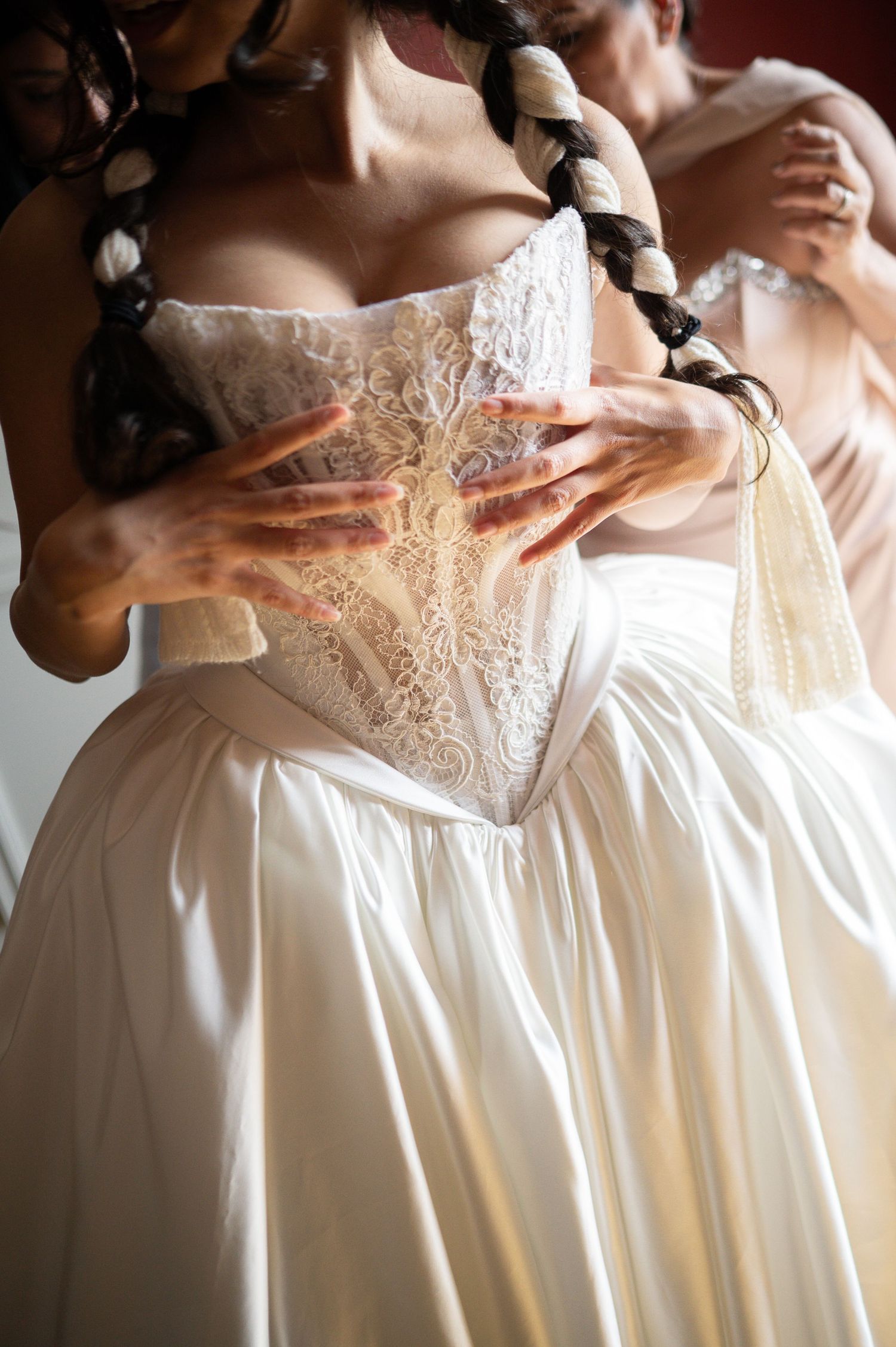 Close-up of bridesmaids adjusting bride's lace corset bodice on voluminous satin ball gown wedding dress.