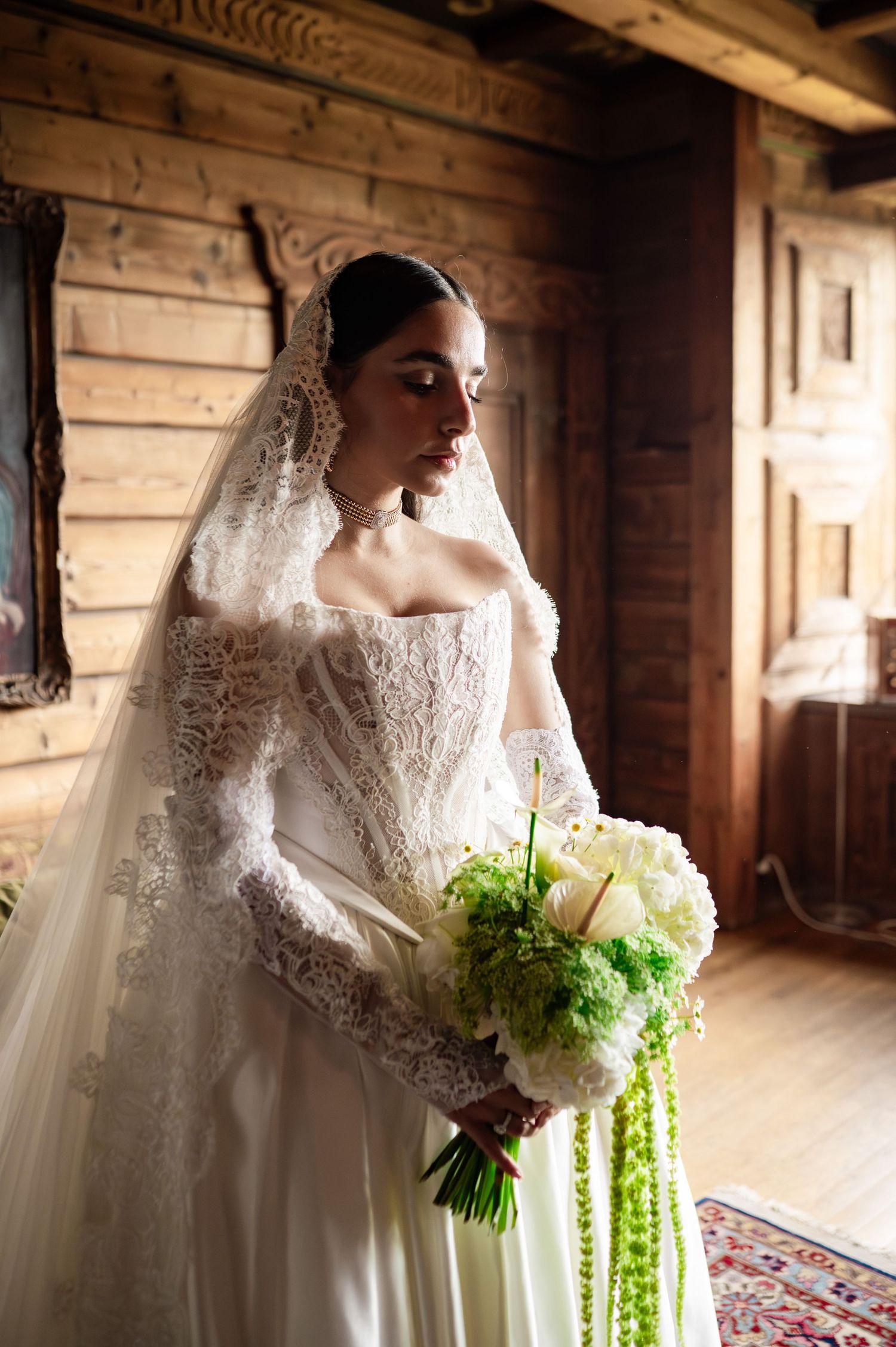 Bride in lace wedding gown with veil holds green and white bouquet in rustic wooden interior.
