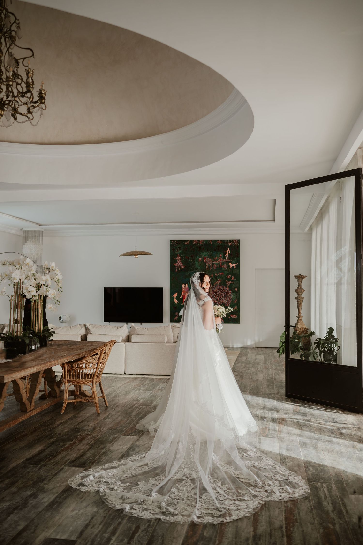 Bride in elegant lace wedding gown with cathedral veil stands in luxurious white interior room.