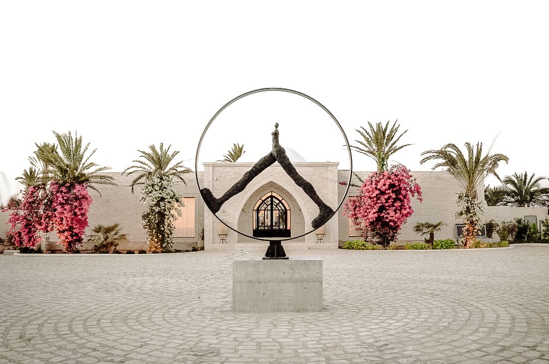 Elegant wedding venue entrance with circular metal sculpture, palm trees, and pink floral arrangements.