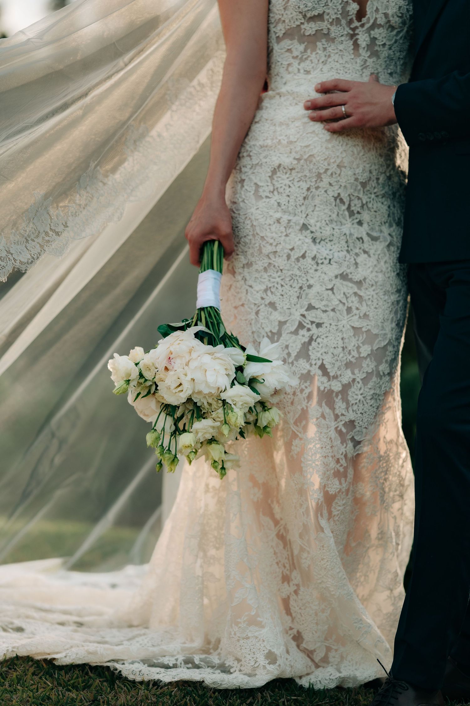 Close-up of bride in intricate lace mermaid gown holding white peony bouquet, veil billowing beside her.