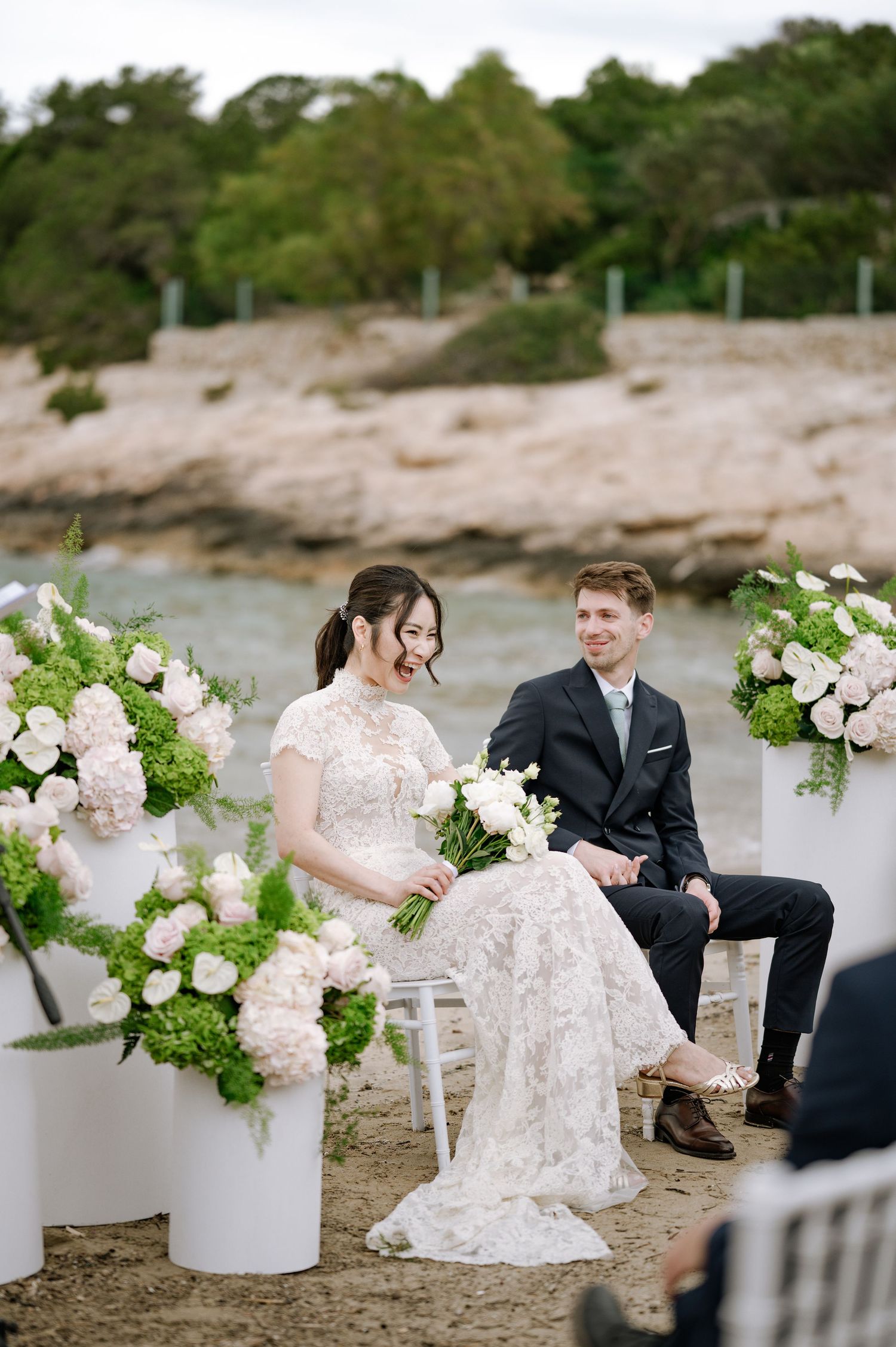 Bride and groom laugh joyfully during beachside wedding ceremony surrounded by white floral arrangements.