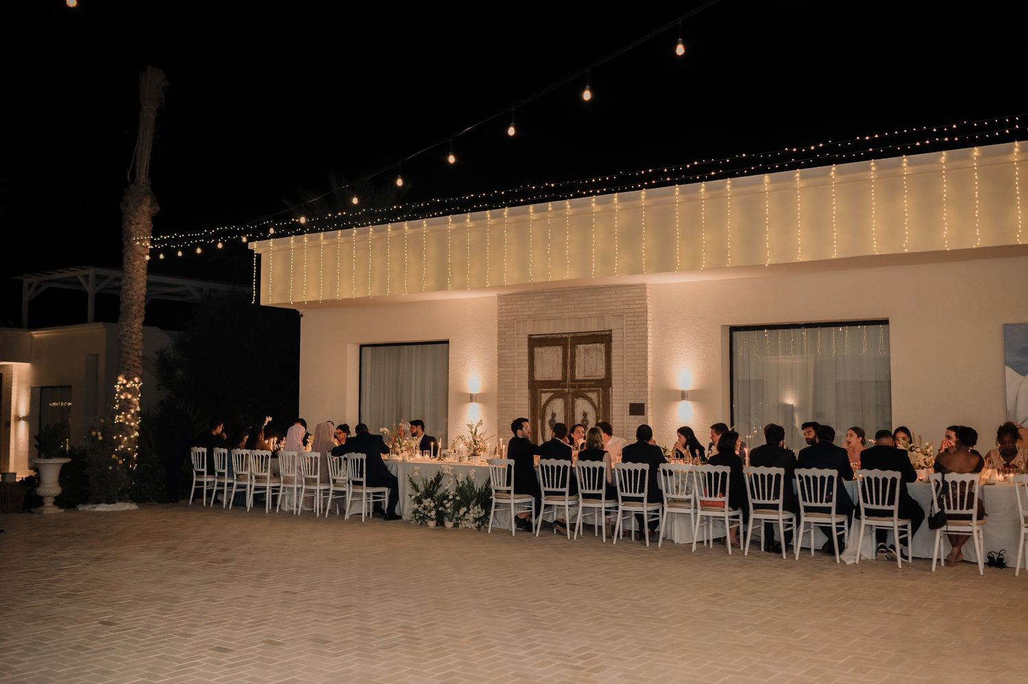 Elegant outdoor wedding reception with long dining table, string lights, and white building backdrop at night.