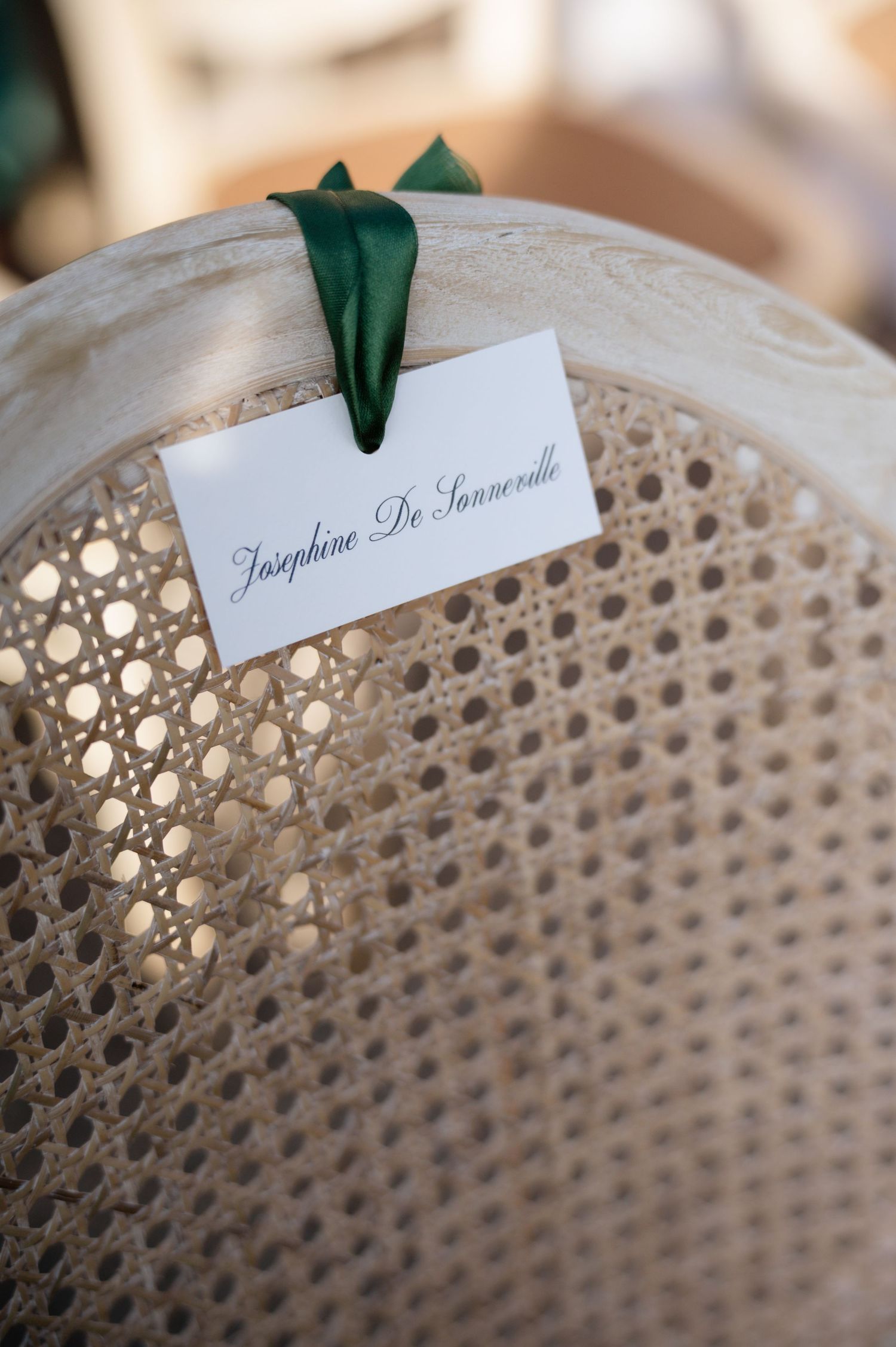 Elegant white place card reading Josephine De Sonneville tied with green ribbon on wicker chair.