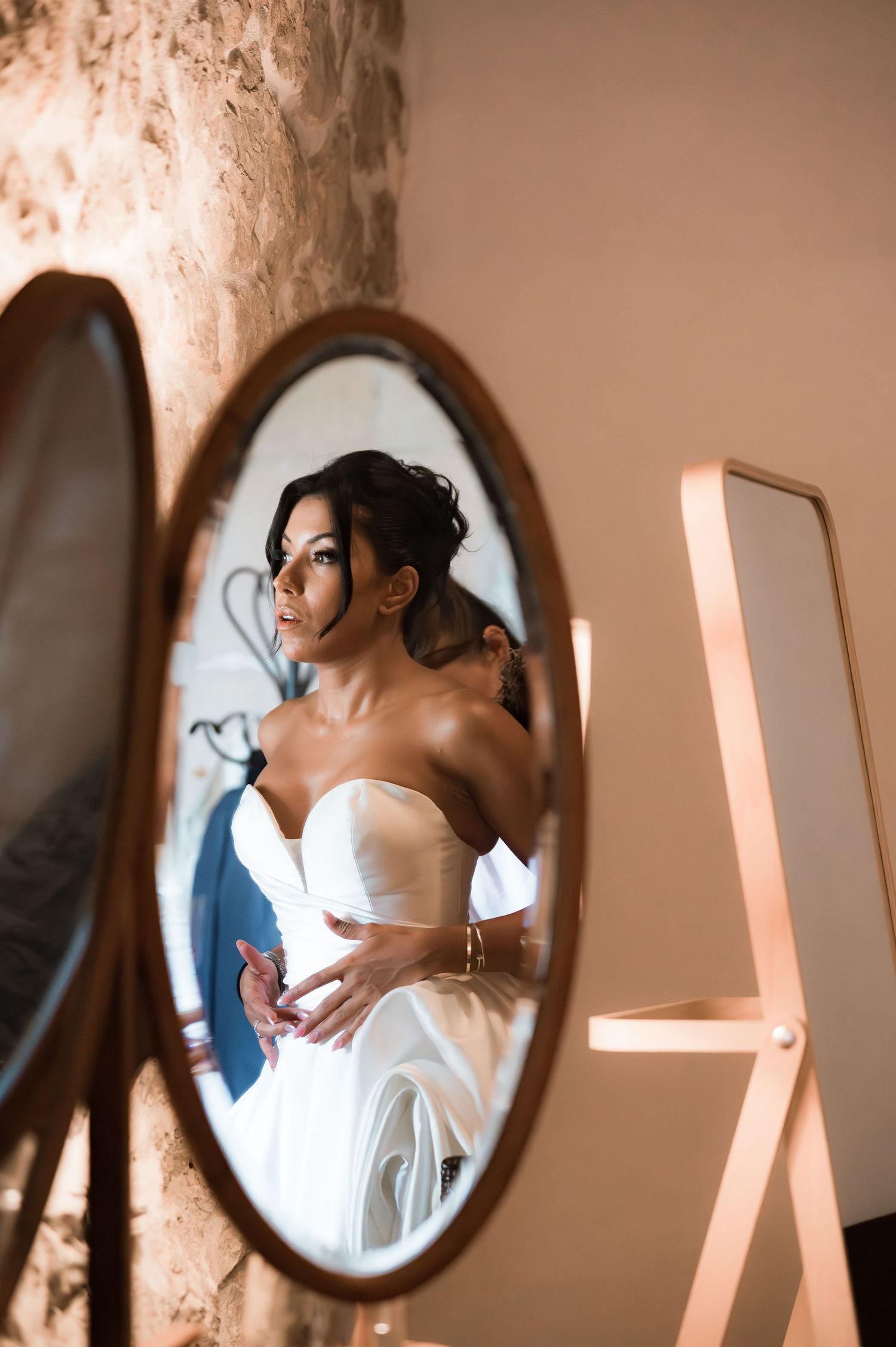 Bride in strapless white wedding gown reflected in oval wooden mirror against rustic stone wall.
