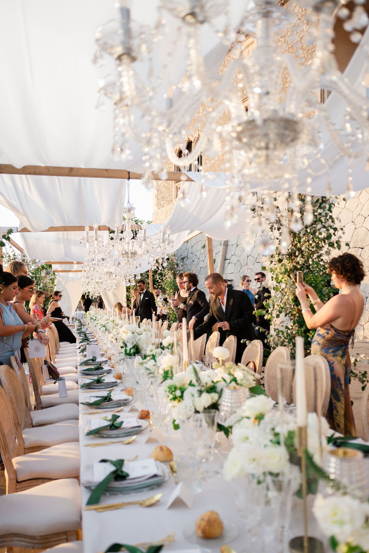 Guests gathered at elegant outdoor wedding reception under crystal chandeliers with white floral table arrangements.