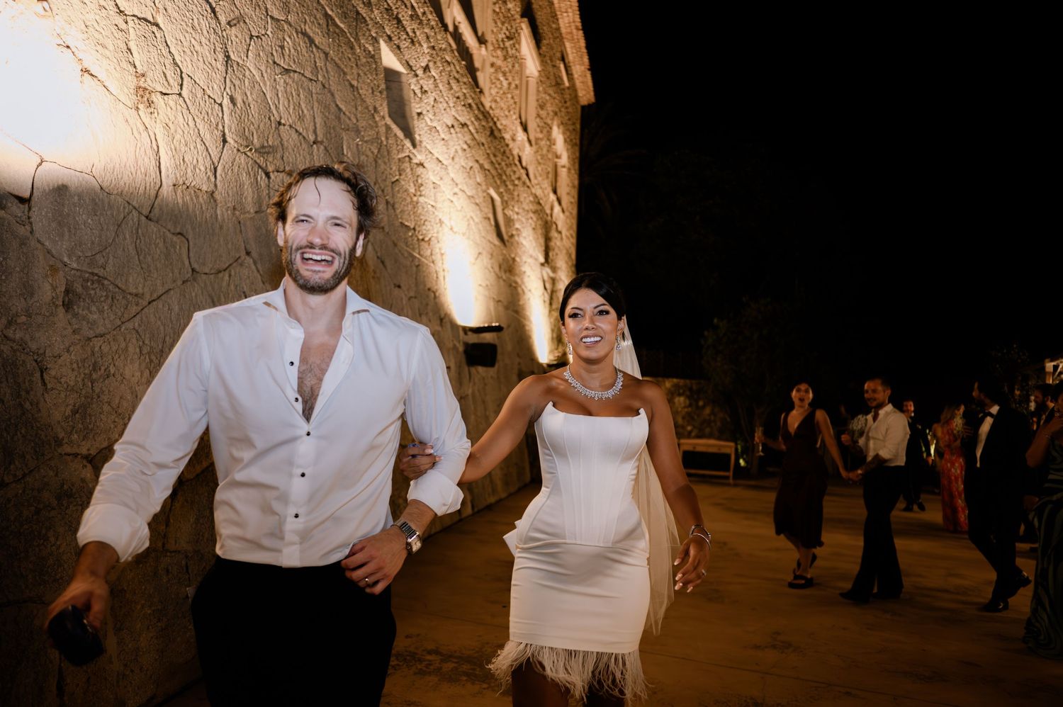 Happy couple in white outfits hold hands and laugh while walking along stone wall at night wedding.