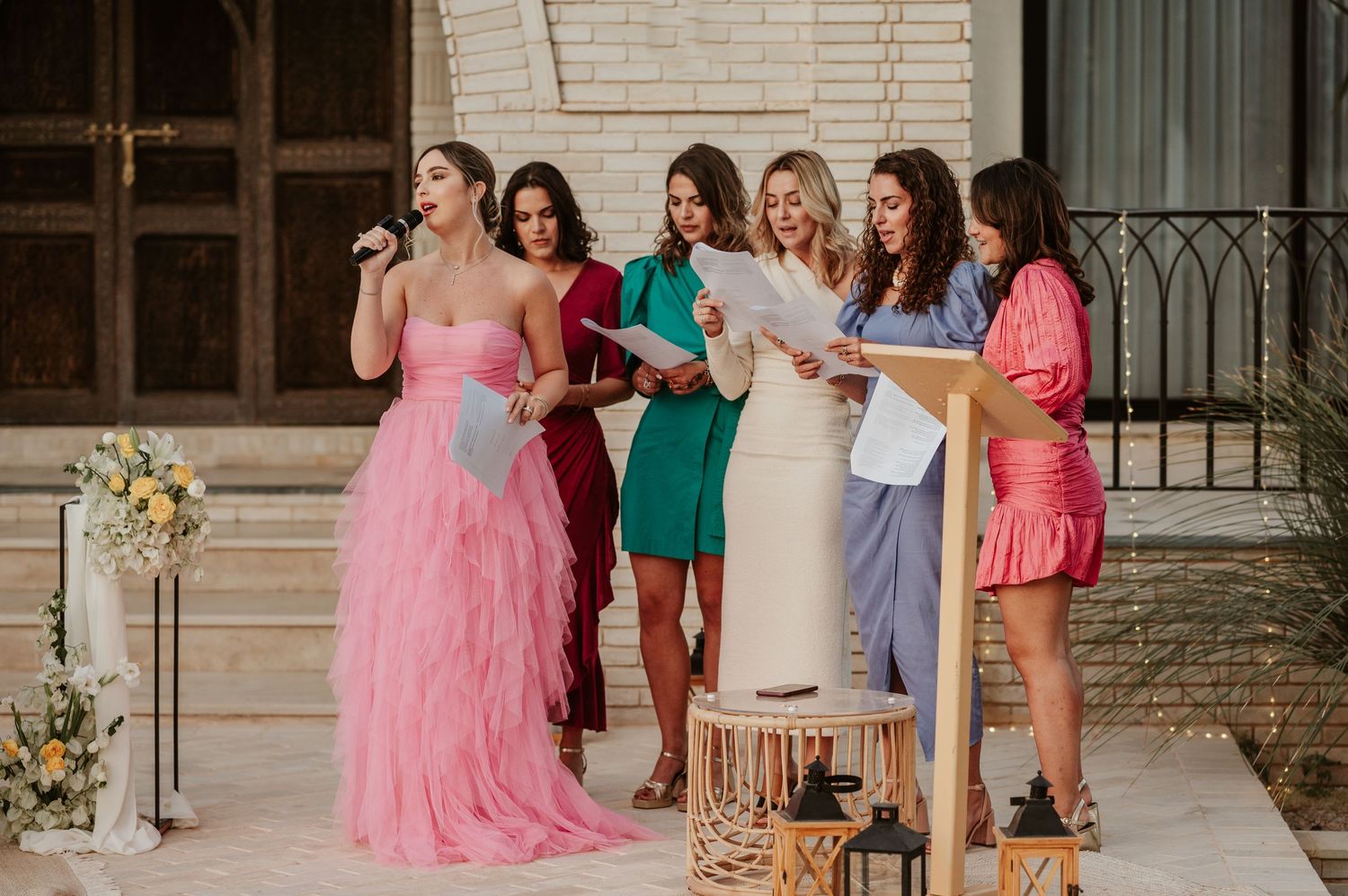 Woman in pink tulle gown sings at microphone with bridesmaids reading along at outdoor wedding ceremony.