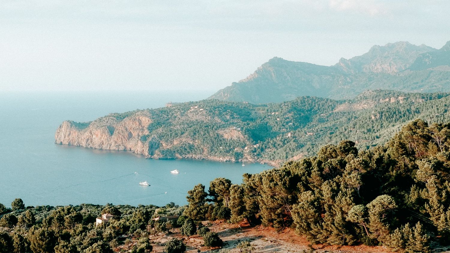 Panoramic aerial view of a lush Mediterranean coastline with deep blue waters, forested hills, and distant mountains.