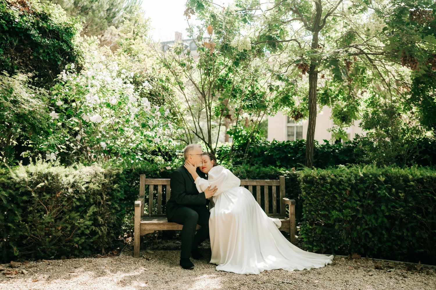 Bride and groom sharing a tender kiss on a wooden bench in a sunlit garden setting.