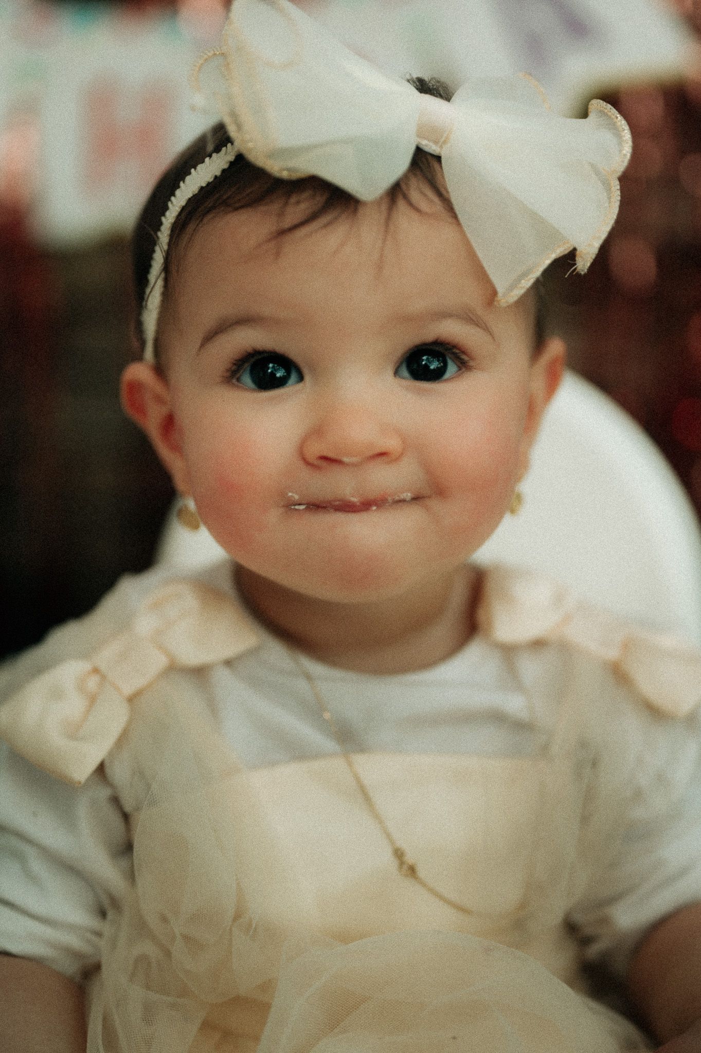 Adorable baby girl with big blue eyes wearing a white bow headband and elegant cream dress smiling sweetly.