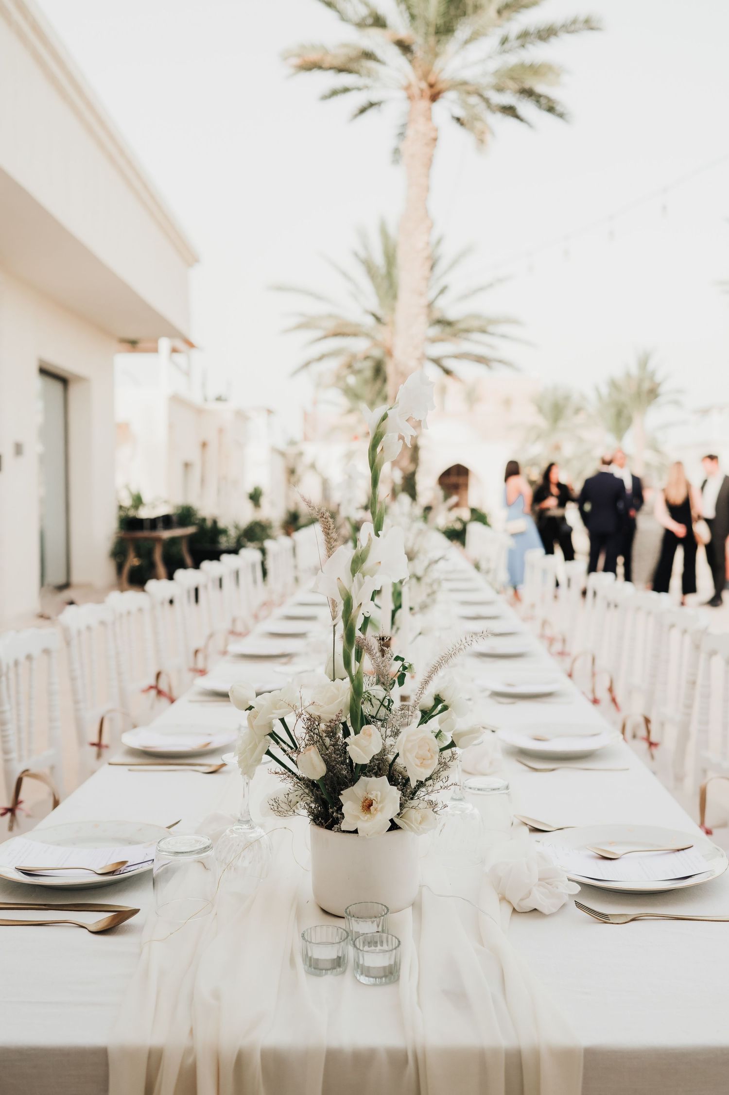 Long outdoor wedding reception table with white floral centerpieces, gold cutlery, and palm trees at dusk.