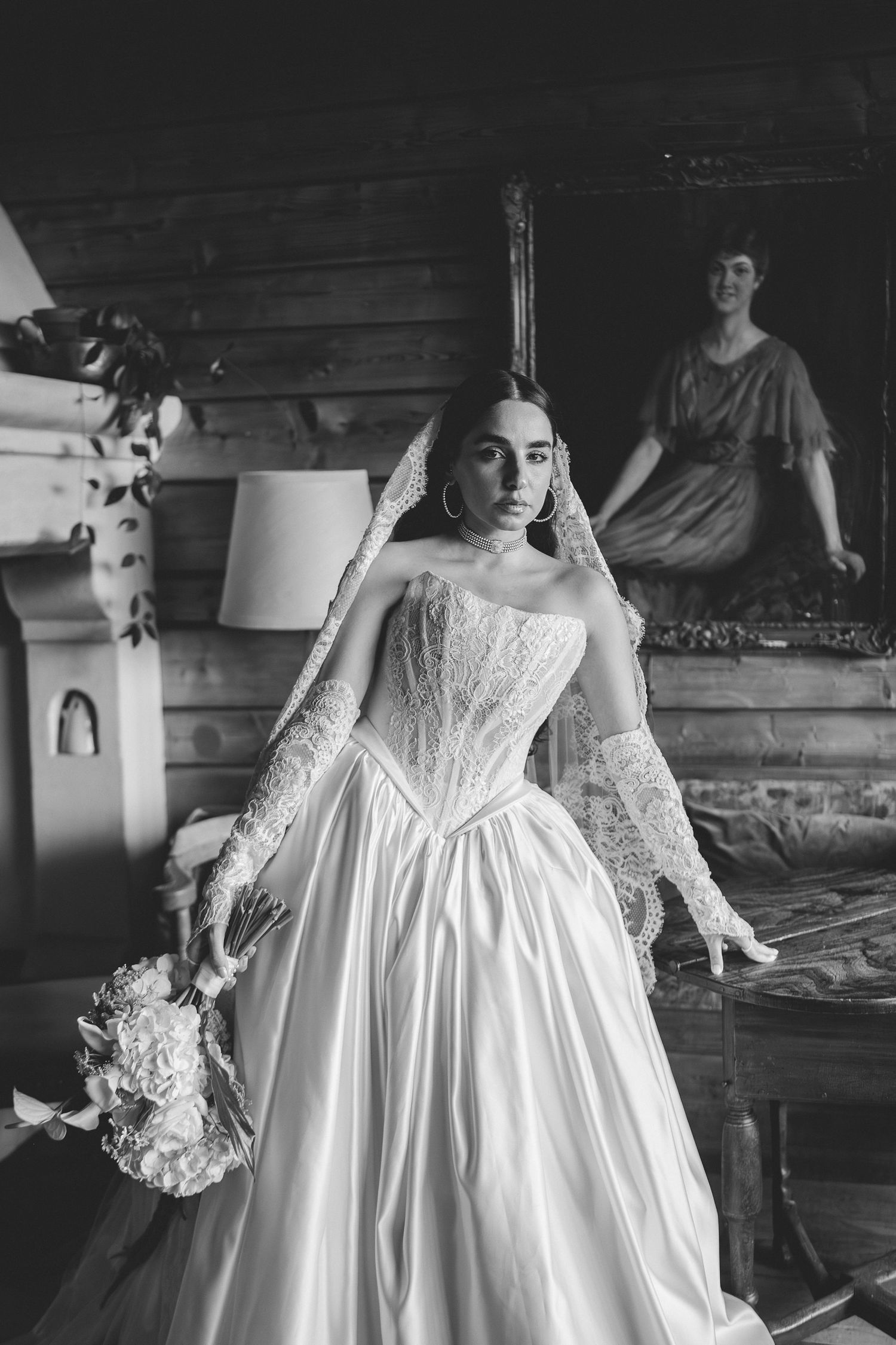 Black and white photo of bride in strapless corset gown with lace gloves holding bouquet indoors.