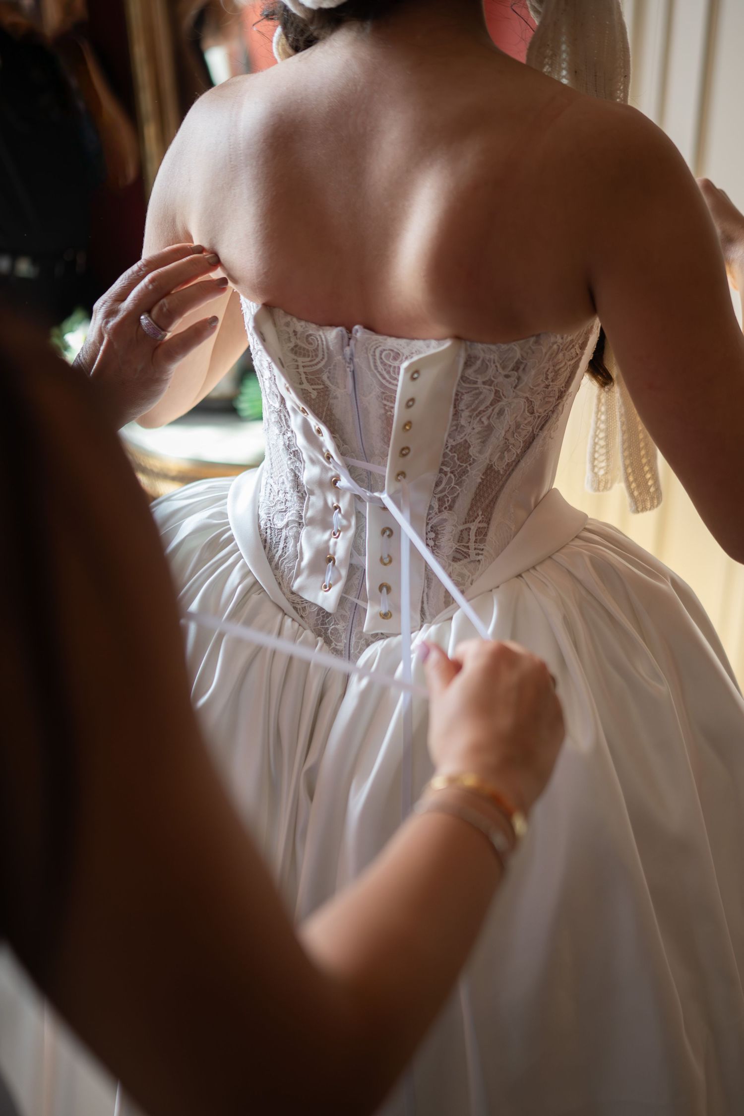 Bride getting corset wedding dress laced up by bridesmaid, showing intricate lace back detail.
