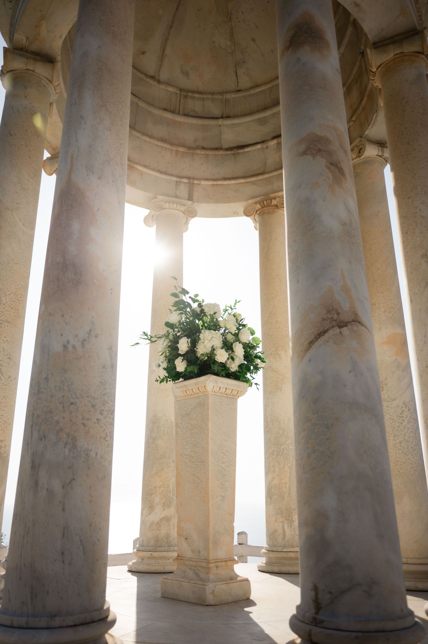 White floral arrangement on a pedestal between marble columns at a wedding venue.