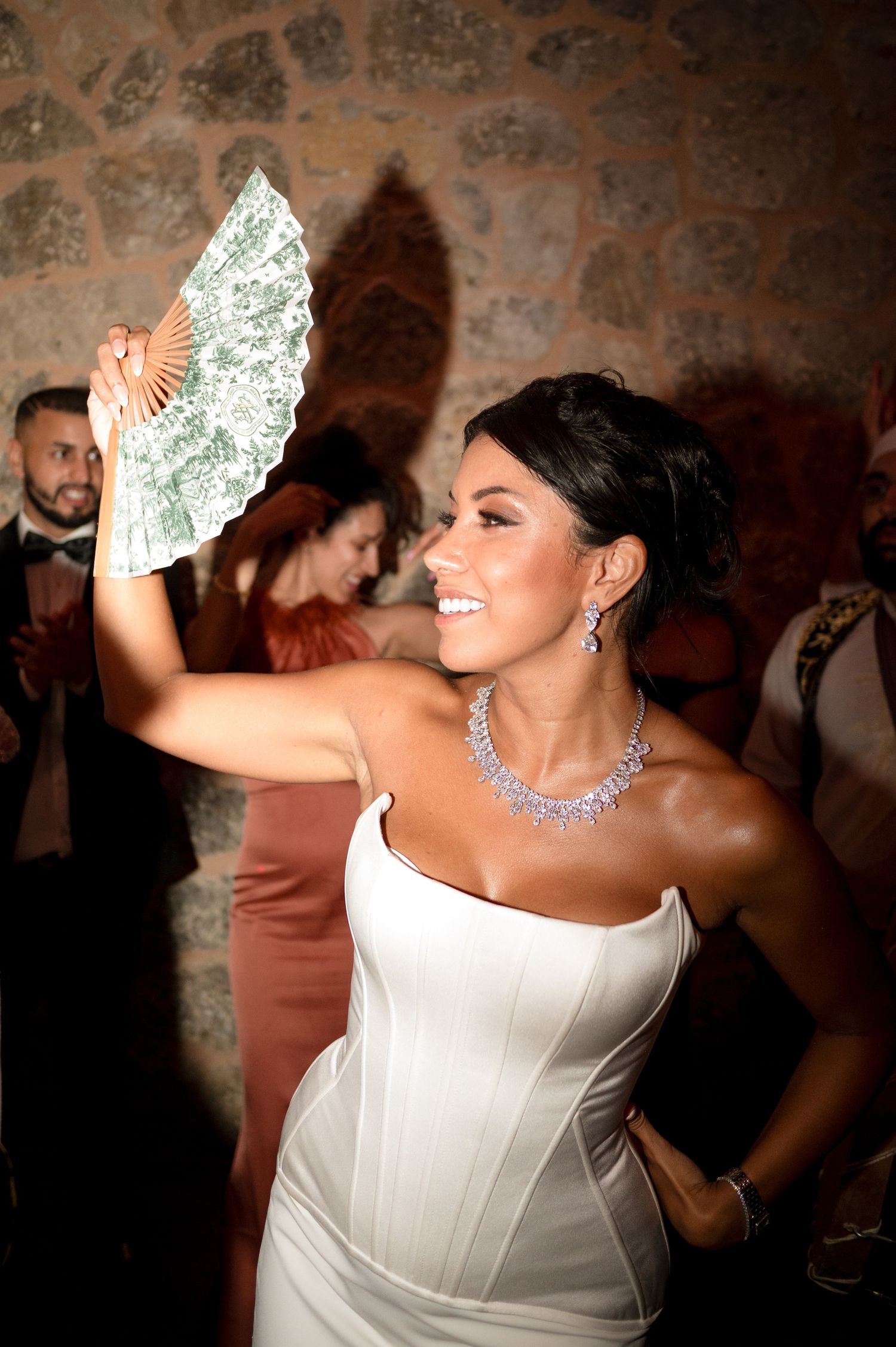 Elegant bride in white strapless gown holding decorative fan at wedding reception.