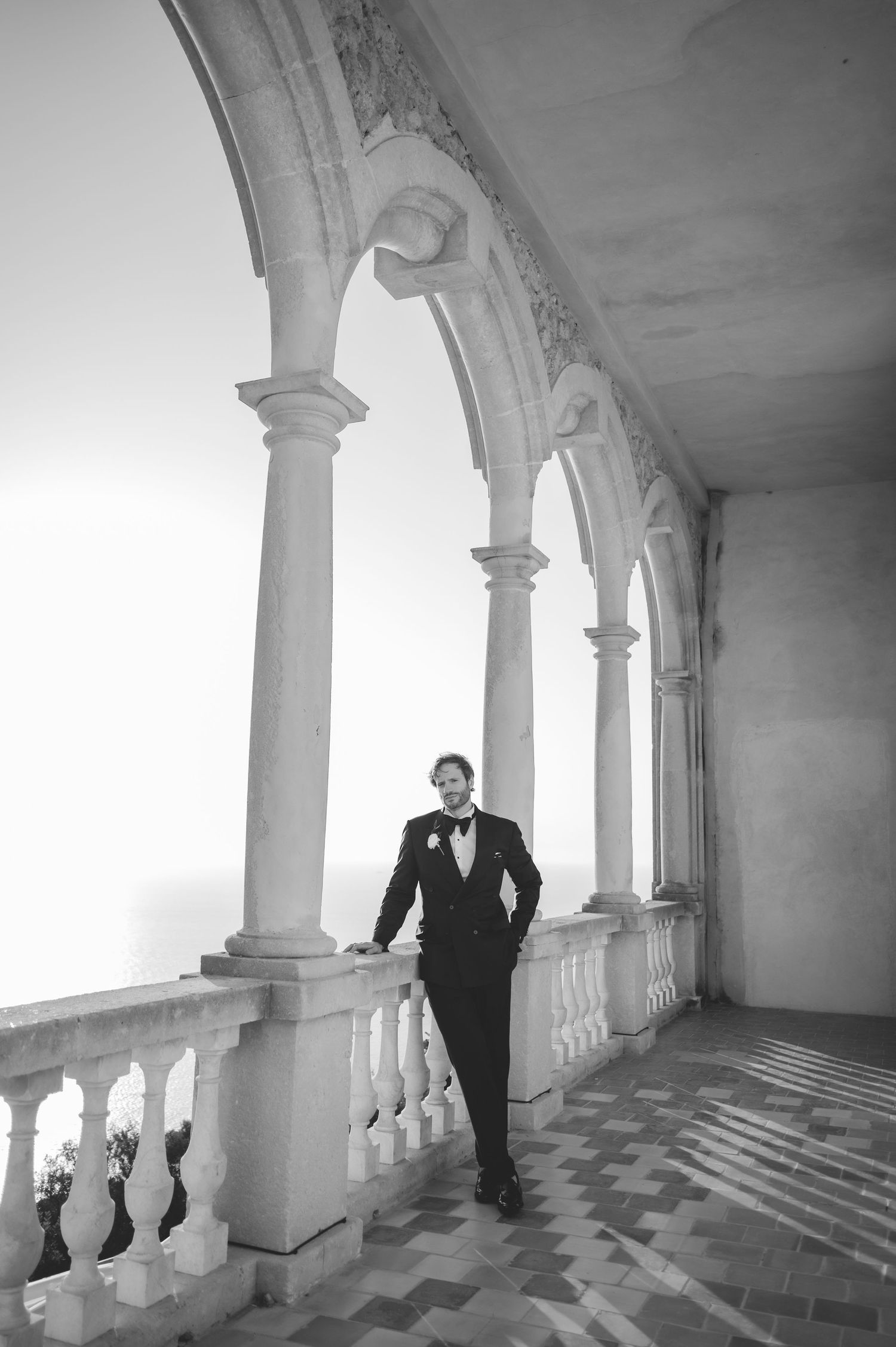 Elegant groom in black tuxedo stands beneath arched stone colonnade in black and white portrait.