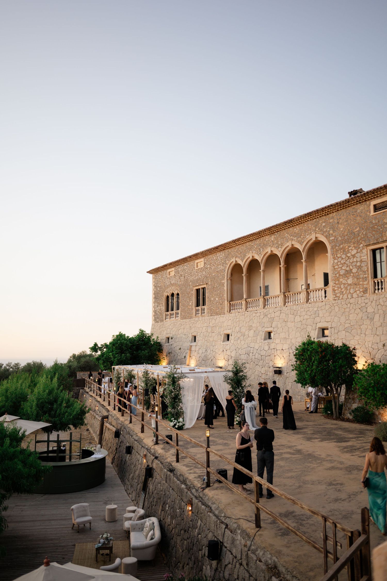 Historic stone villa wedding venue at dusk with guests mingling on terrace surrounded by lush greenery.