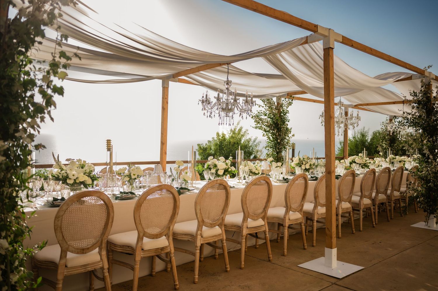 Outdoor wedding reception with long dining table, Louis chairs, white draped canopy, chandeliers, and ocean view backdrop.