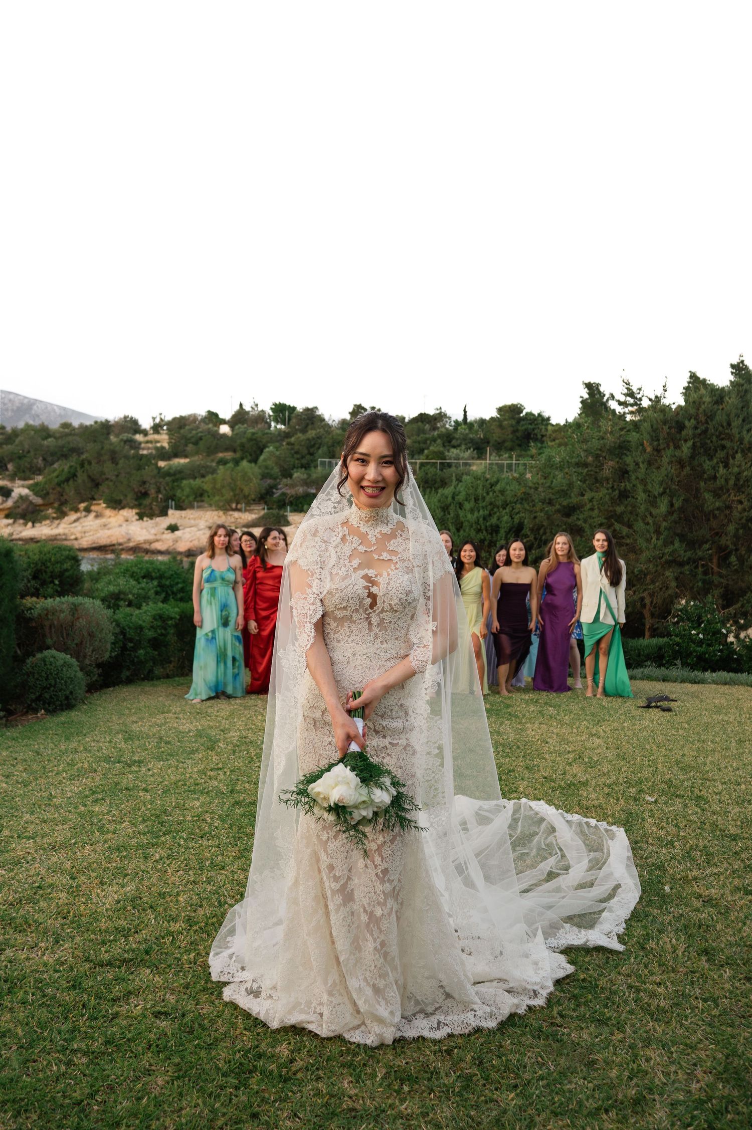 Bride in lace wedding gown and veil holds bouquet with colorful bridesmaids standing behind on green lawn.