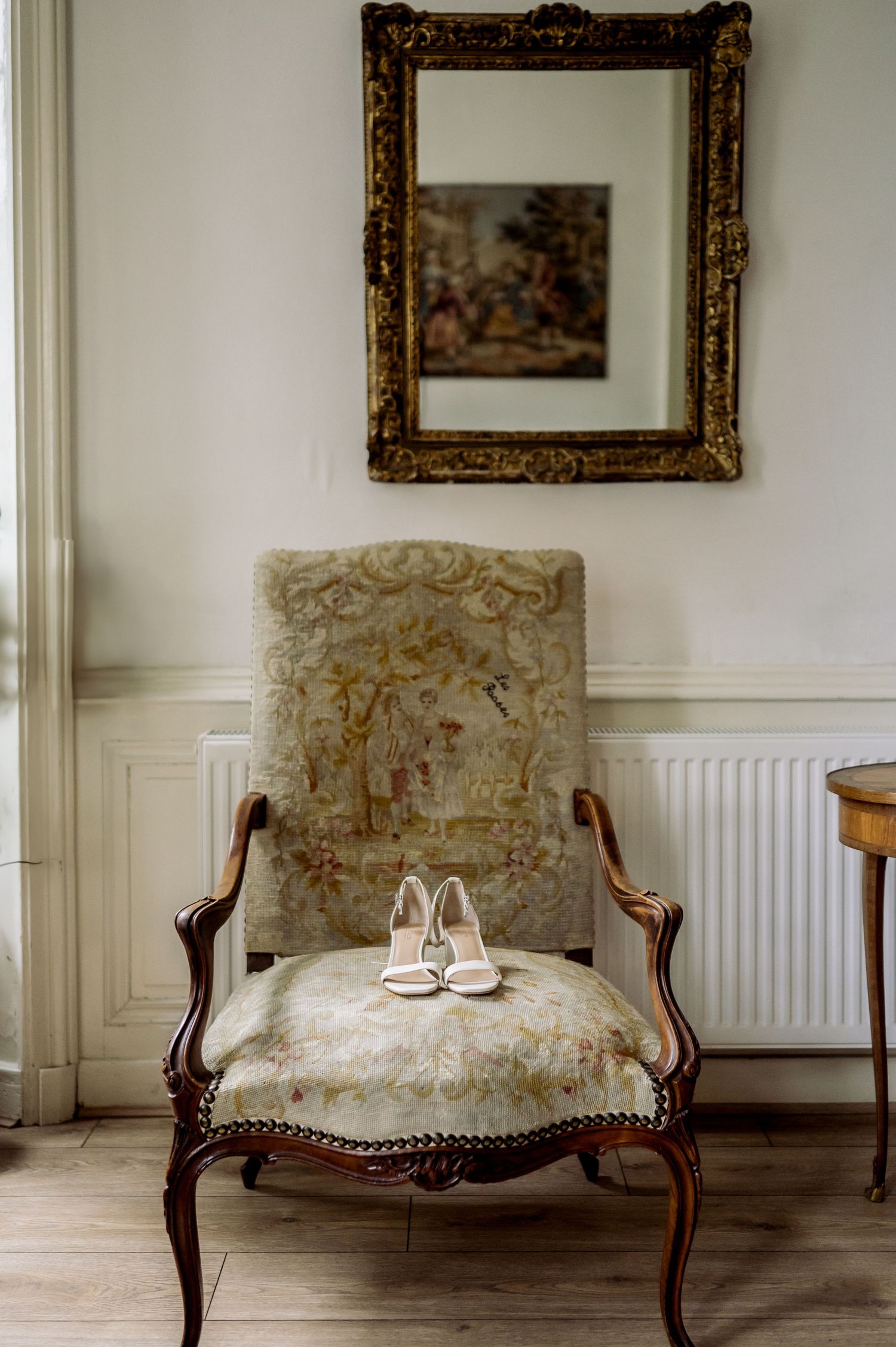 Vintage French armchair with ornate gold mirror above holding white bridal heels in an elegant room.