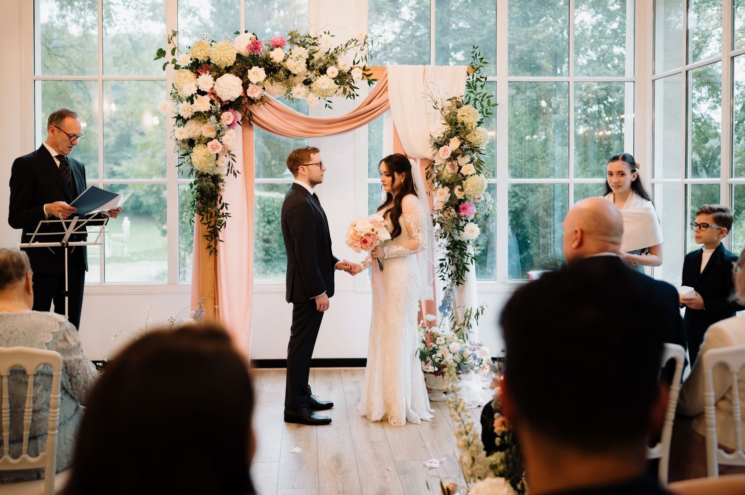 Bride and groom stand at a floral wedding arch during their indoor ceremony with seated guests.