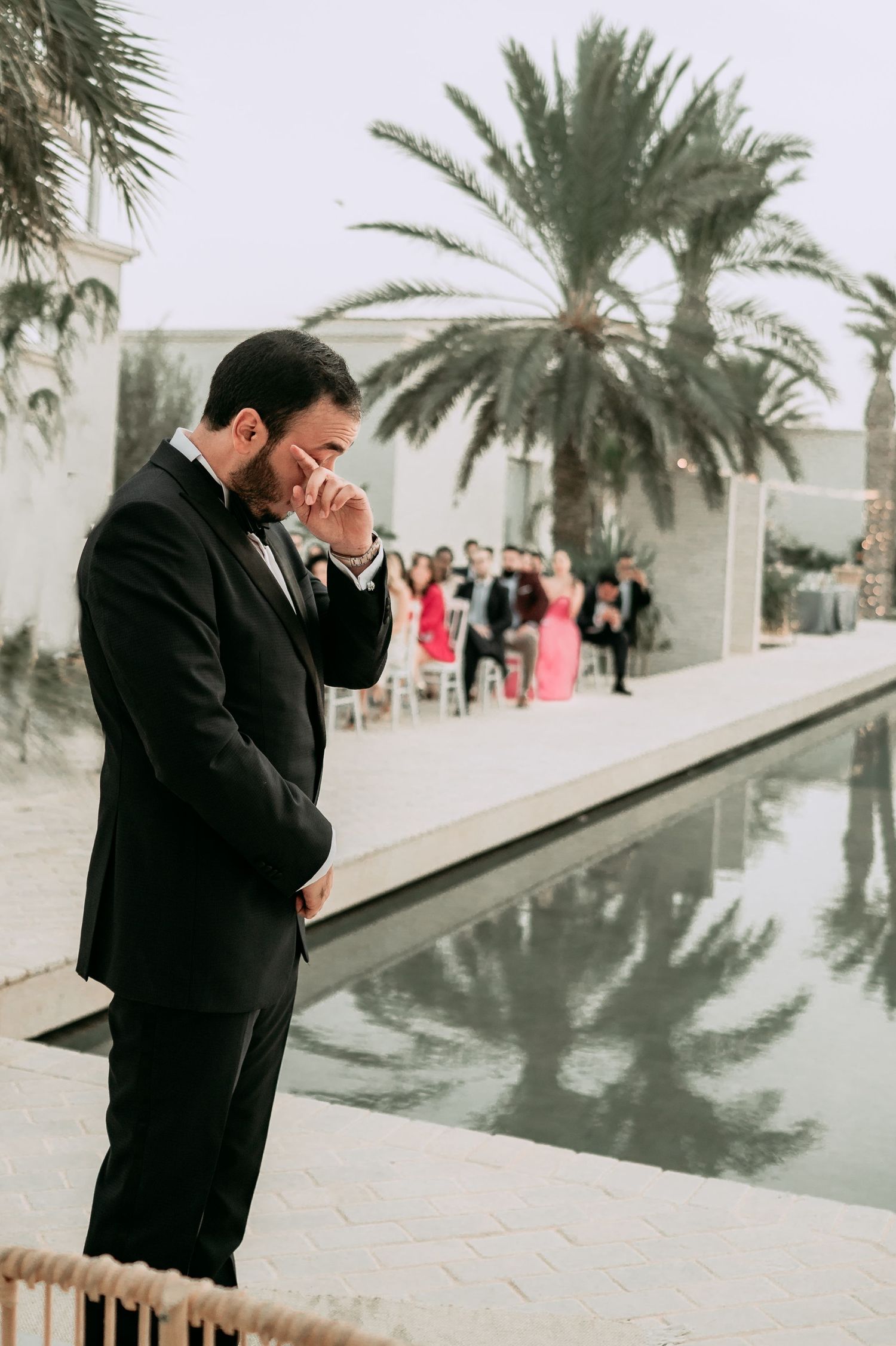 Emotional groom in black tuxedo wipes tears at outdoor wedding ceremony beside a serene reflecting pool.