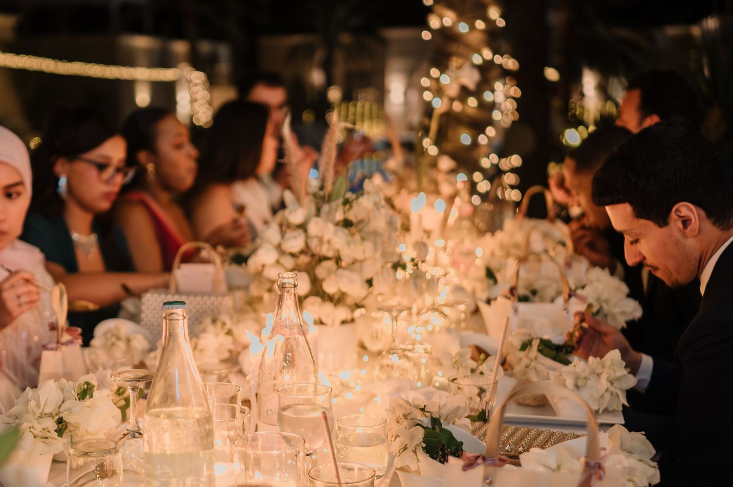 Guests seated at candlelit wedding reception table adorned with white floral arrangements and string lights.