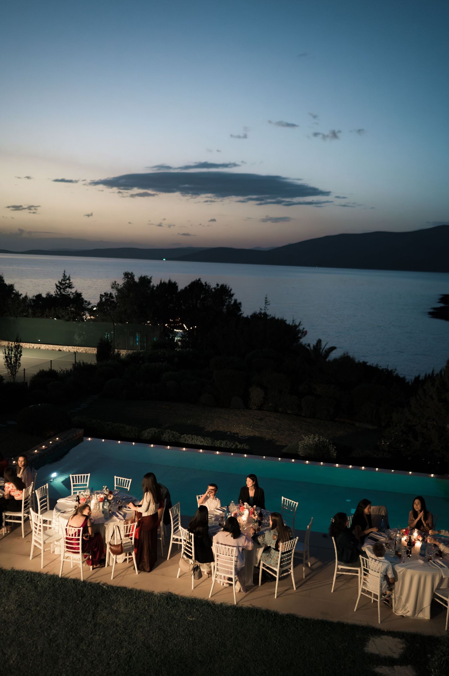 Outdoor wedding reception dinner by illuminated pool at sunset with sea view.