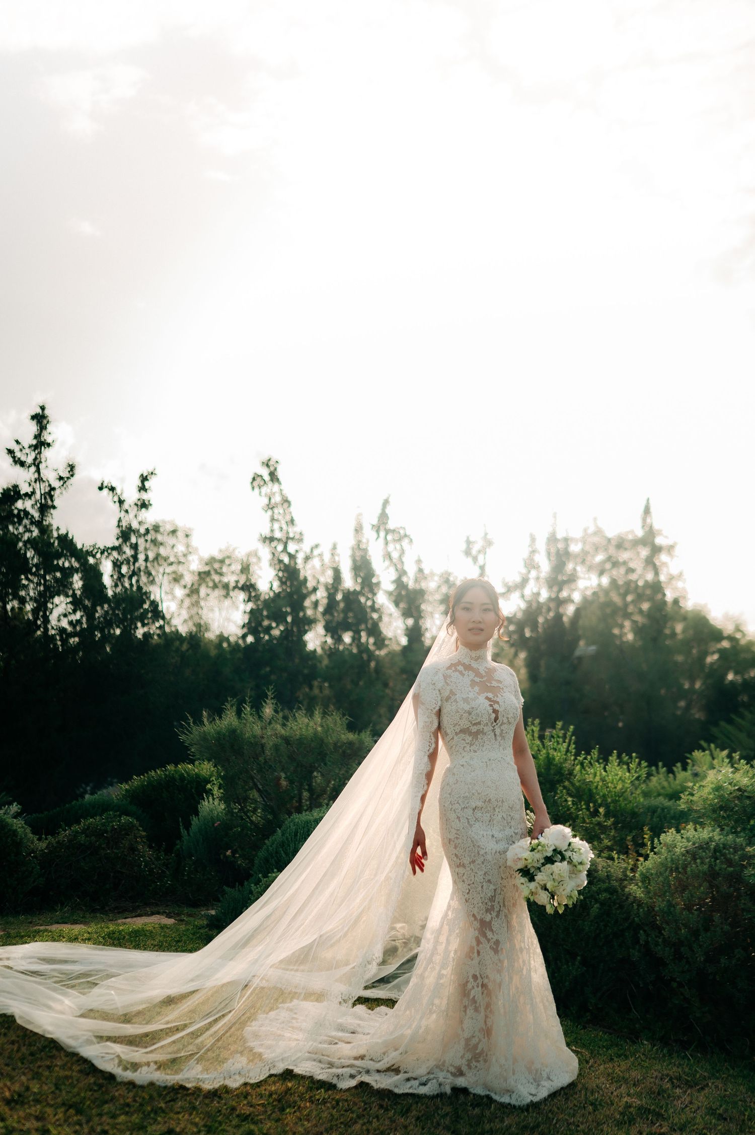 Elegant bride in high-neck lace gown with cathedral veil standing in sunlit garden holding white bouquet.