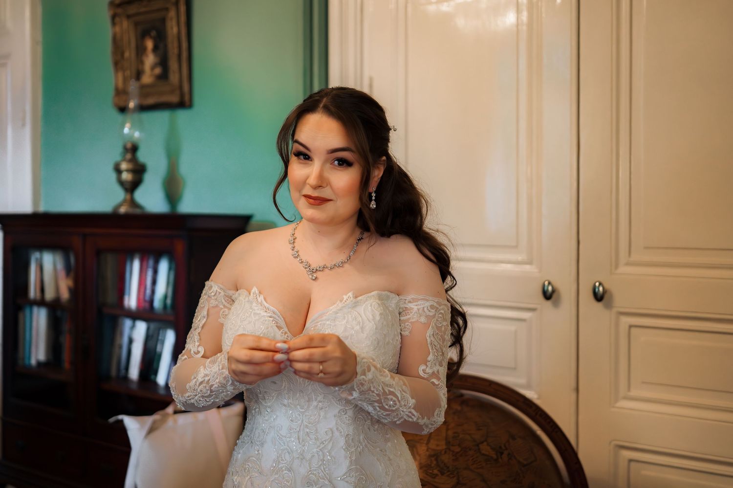 Bride in long-sleeve lace wedding gown fastening jewelry in a elegantly decorated room.