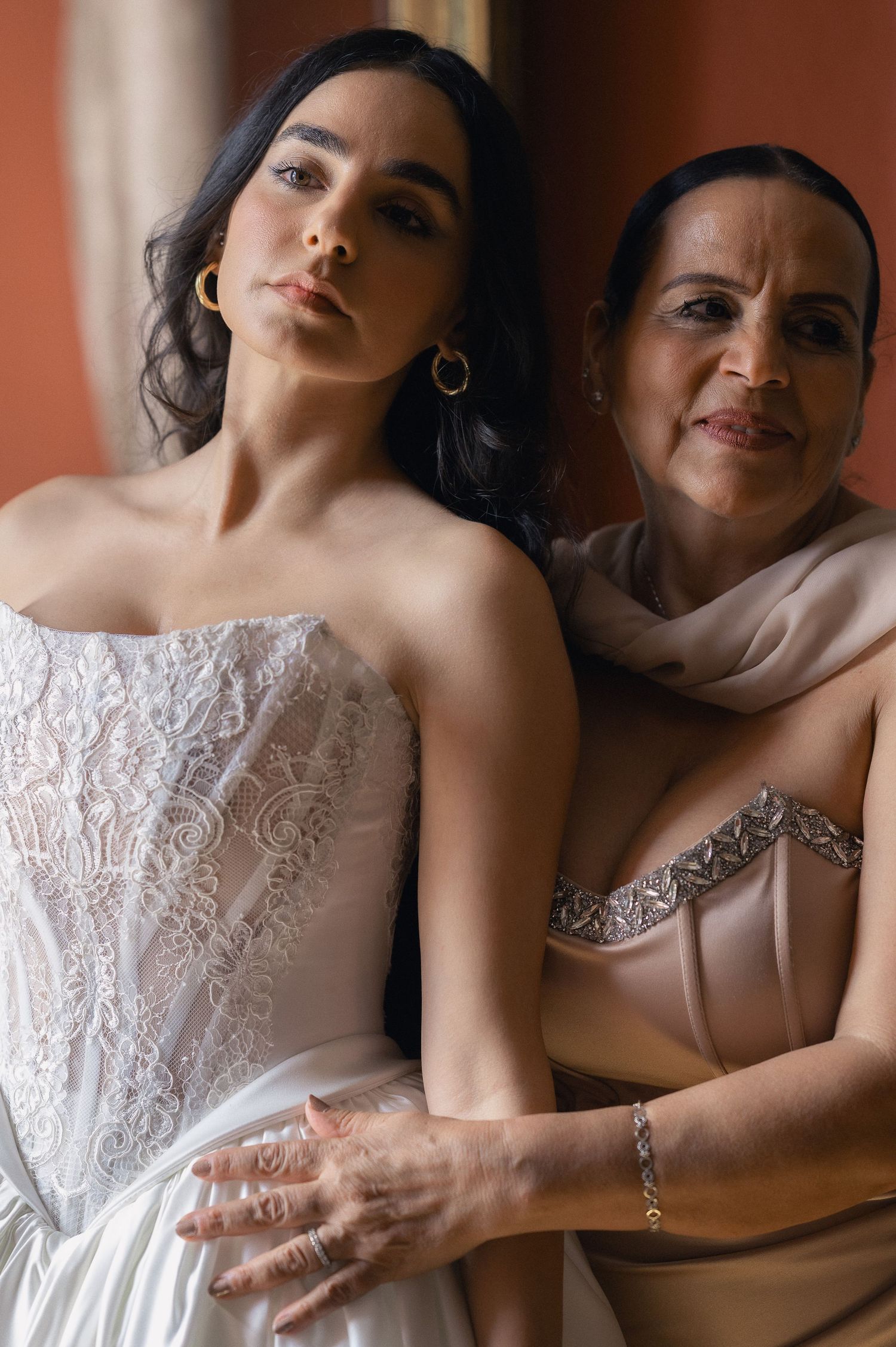 Bride in strapless white lace wedding gown posing with older woman in beige dress in a warm-toned room.