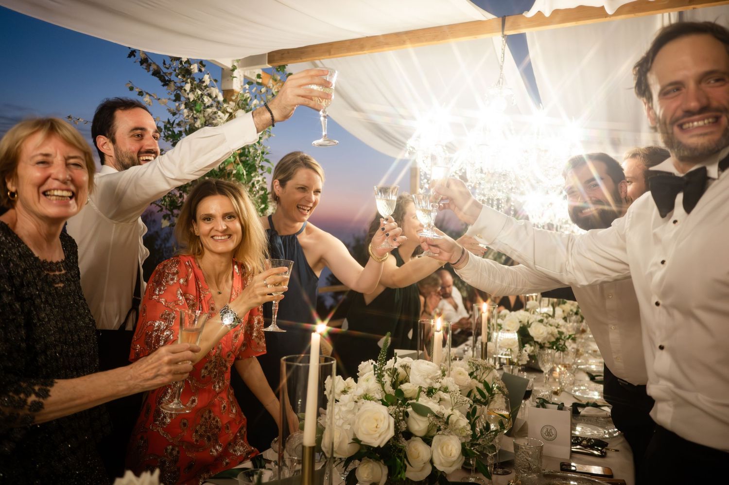 Joyful wedding guests raising champagne glasses in a toast surrounded by white floral centerpieces and sparklers.