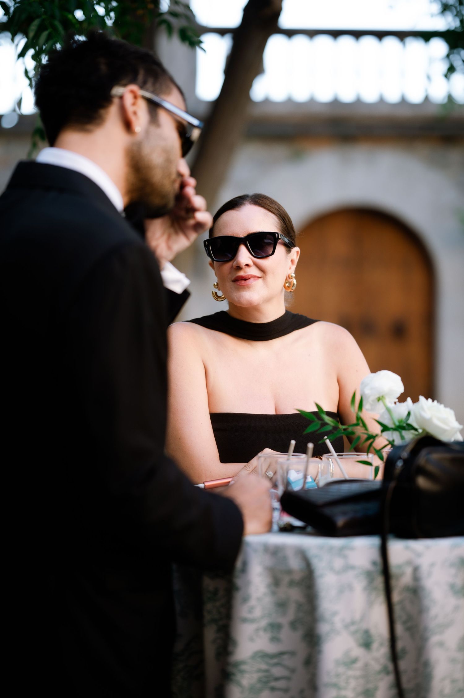 Stylish wedding guest in black strapless dress and sunglasses at outdoor registration table.