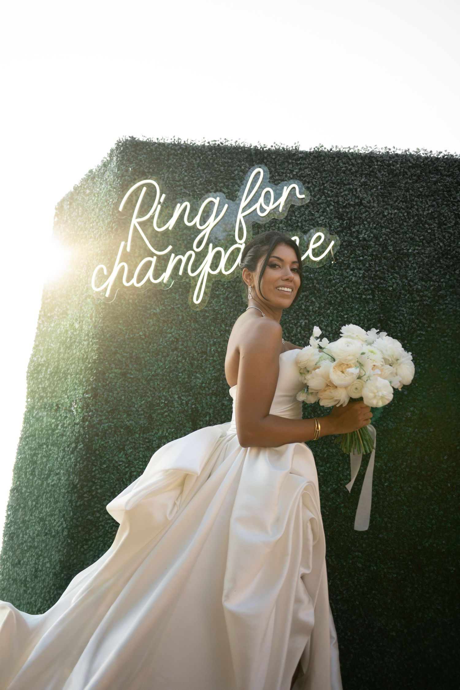 Radiant bride in strapless white ball gown holding white bouquet in front of Ring for Champagne neon sign.