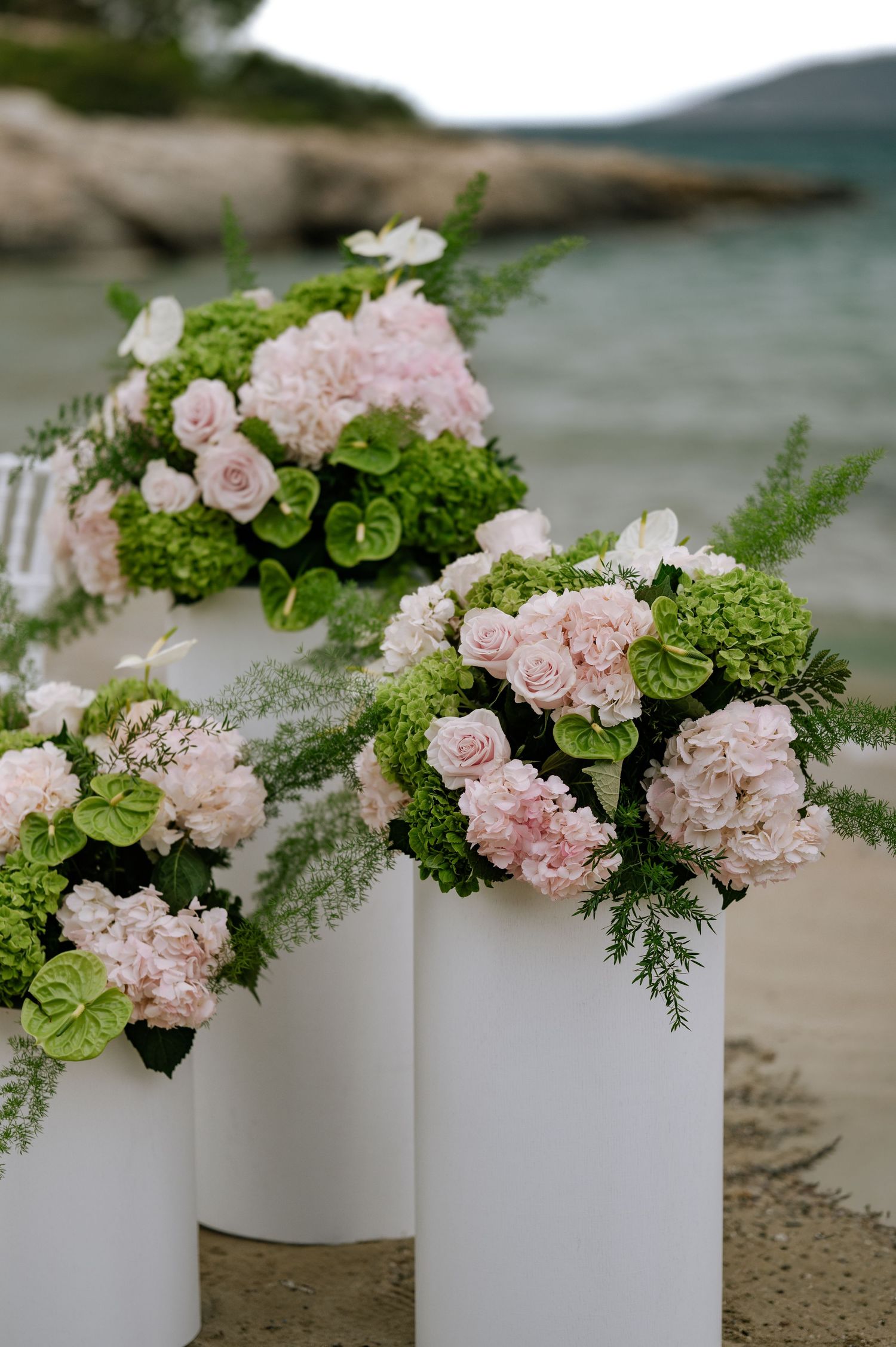 Elegant wedding floral arrangements featuring pink roses, hydrangeas, and green foliage on white cylindrical pedestals by the sea.