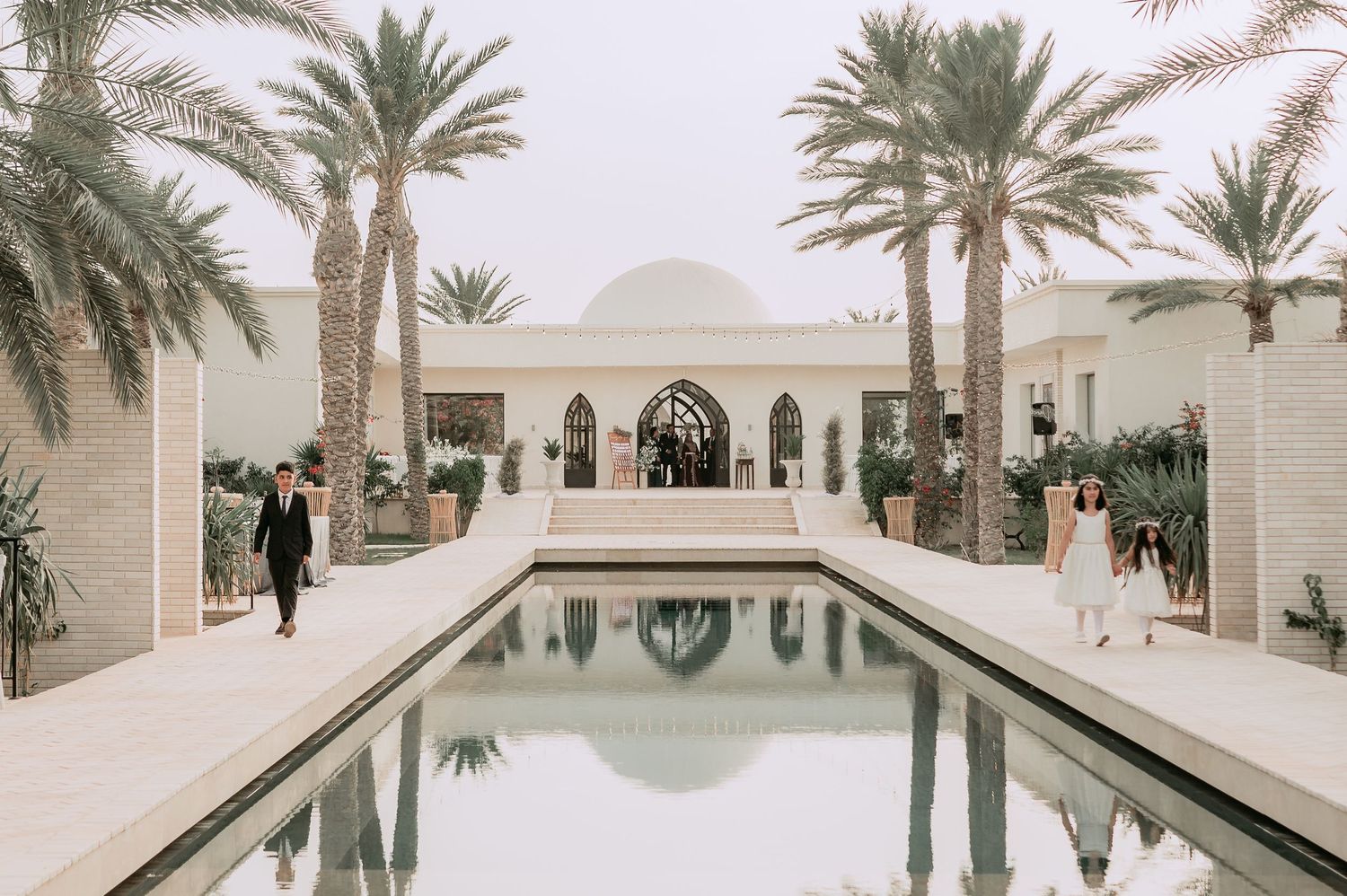 Elegant wedding venue with long reflecting pool, white domed building, and tall palm trees in a desert setting.