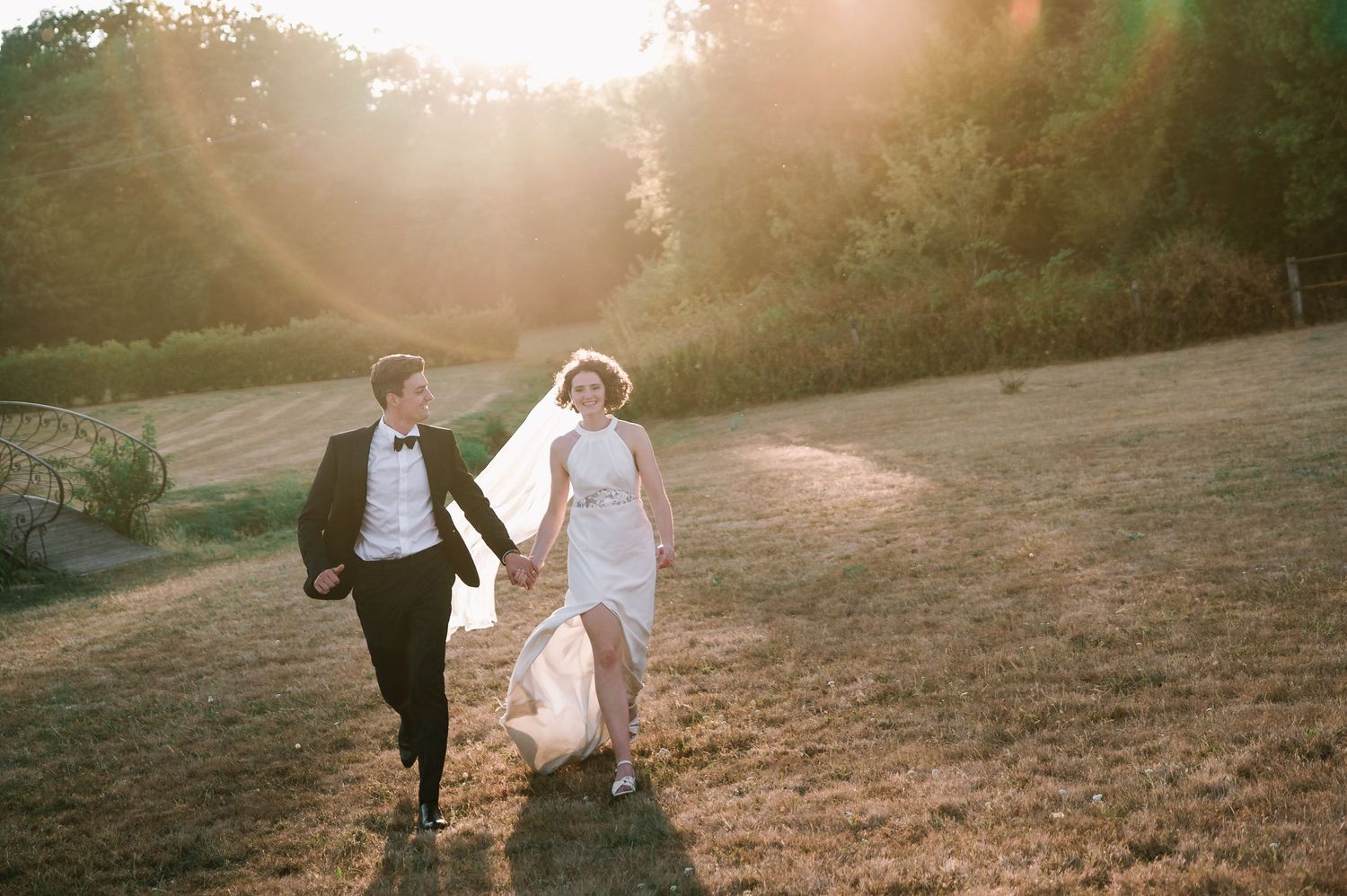 Joyful bride and groom running hand in hand through a golden sunlit meadow on their wedding day.